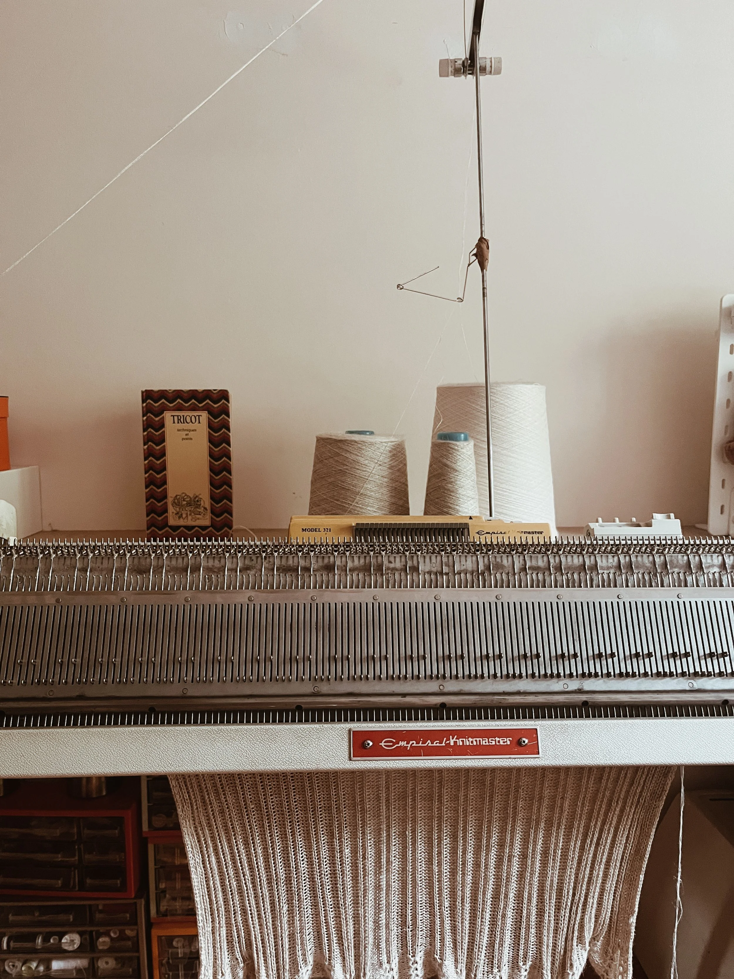 A vintage knitting machine with a partially knitted piece hanging down, yarn spools in the background, and books on the shelf behind.