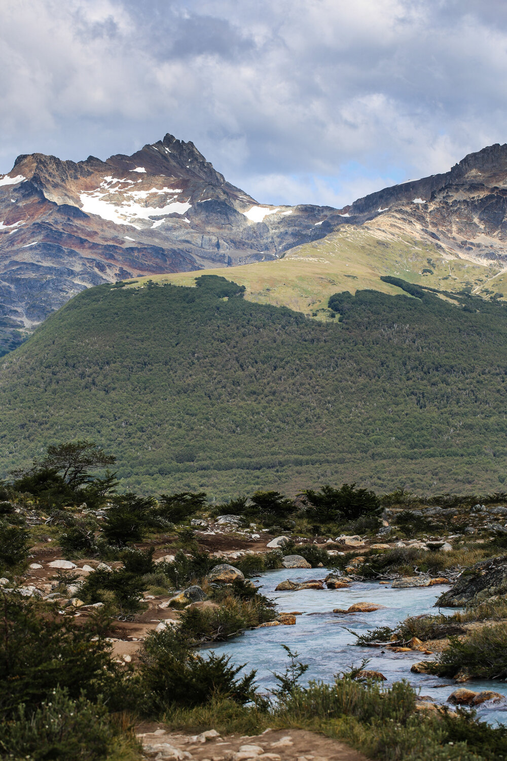 Mountain landscape with a river and green hills, under a cloudy blue sky.