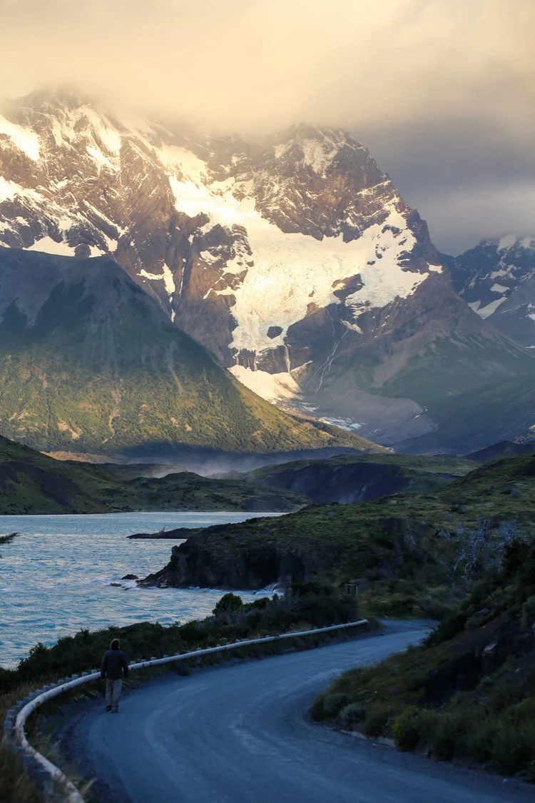 Scenic landscape with mountains, glacier, winding road, and a lone figure walking, under a cloudy sky.