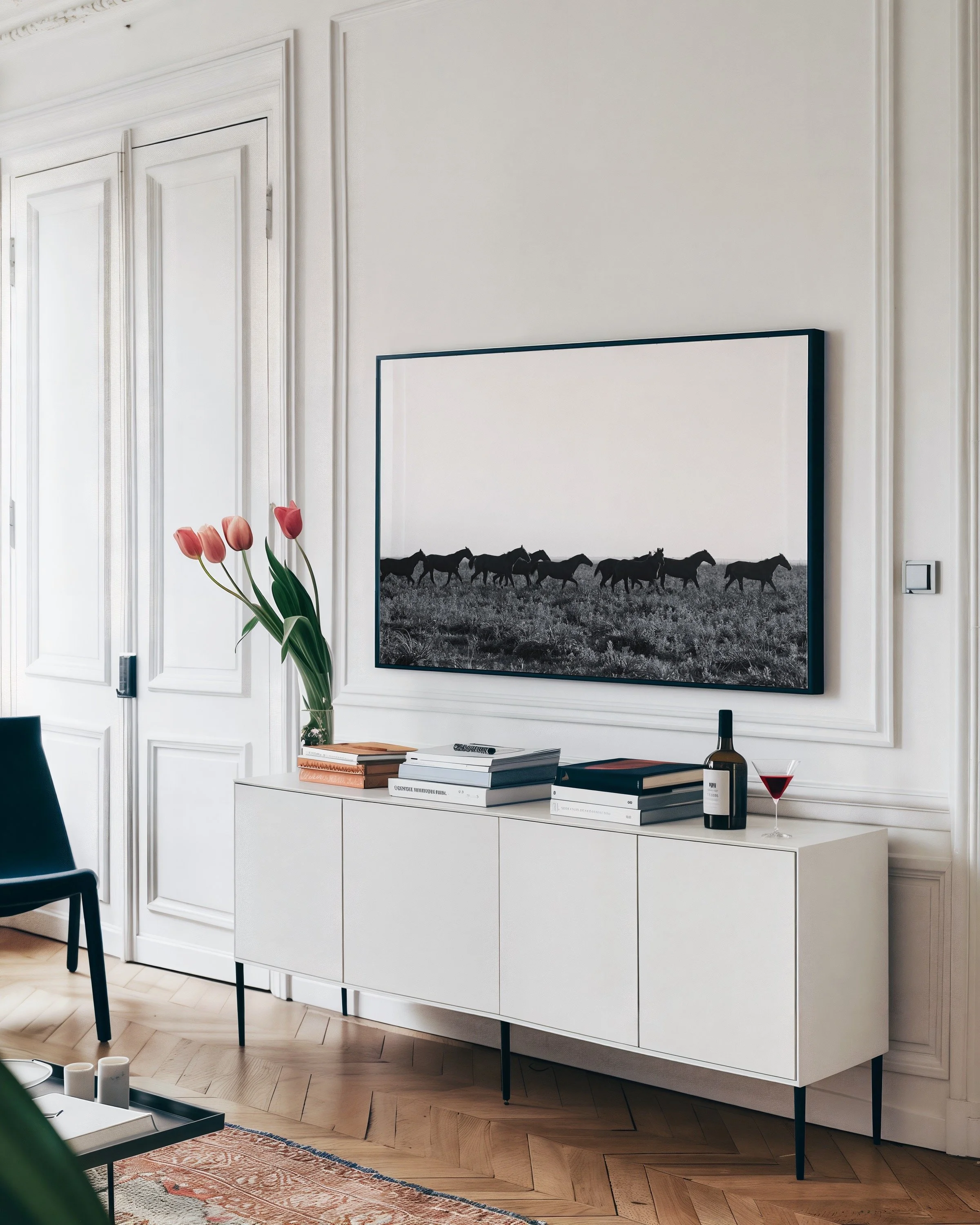 Modern living room interior with white cabinet, framed black and white horse photograph, books, and wine bottle on top, pink tulips in vase, and parquet flooring.