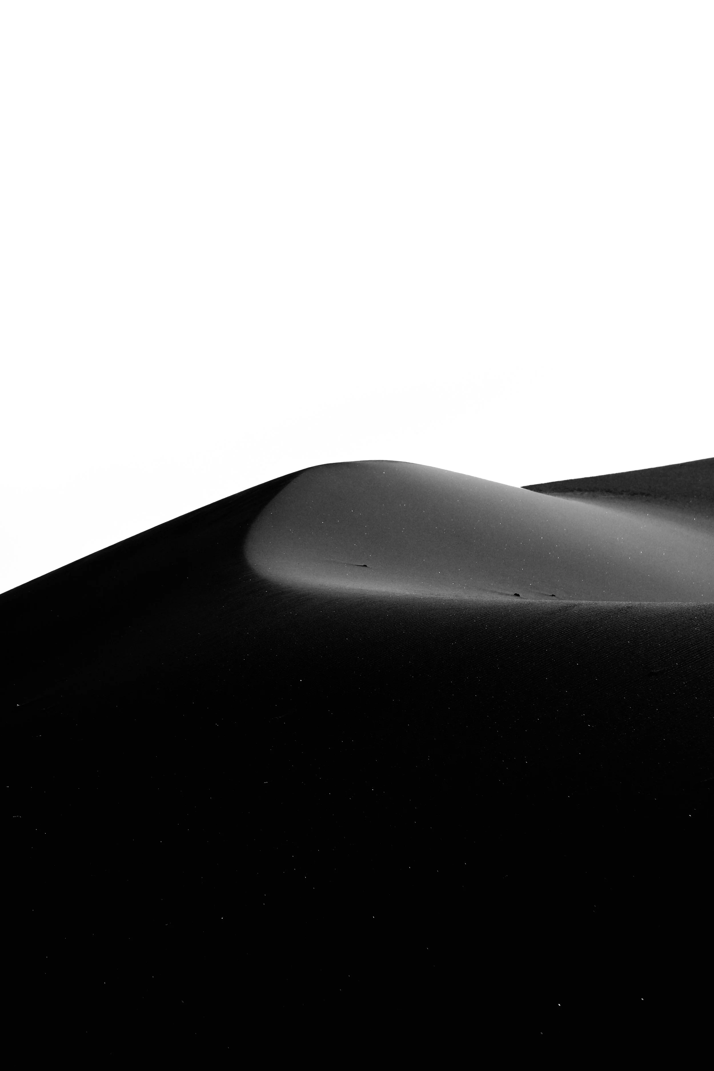 Black and white photograph of a sand dune with a smooth curved surface and shadow contrast against a white sky.