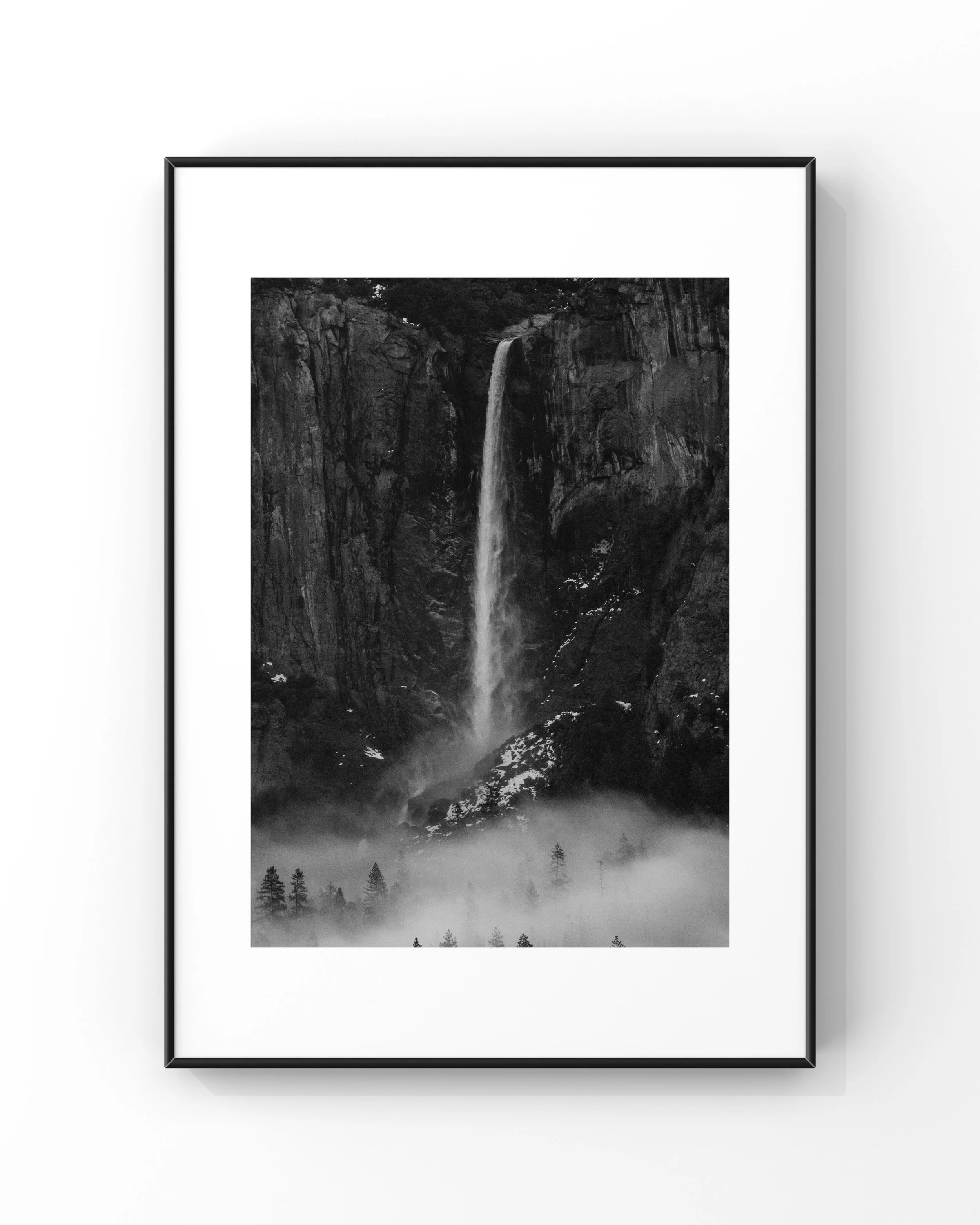 Black and white photo of a waterfall in a mountainous landscape, surrounded by trees and mist, in a framed display.