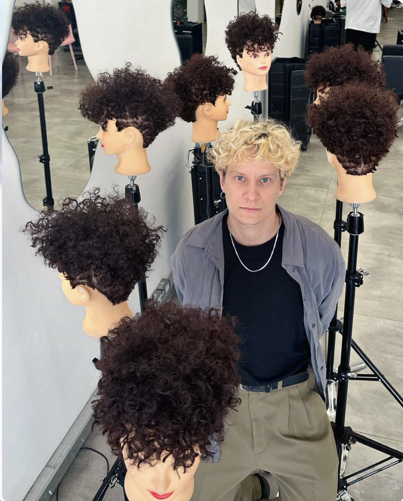 A person with bleached blonde curly hair surrounded by multiple wigs with black curly hair on mannequin heads in a salon.