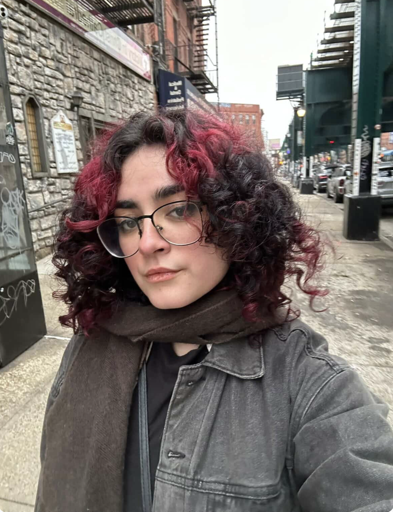 A colorist with curly hair dyed pink and black, wearing glasses, a leather jacket, and a scarf, taking a selfie on a city sidewalk with parked cars and buildings in the background.
