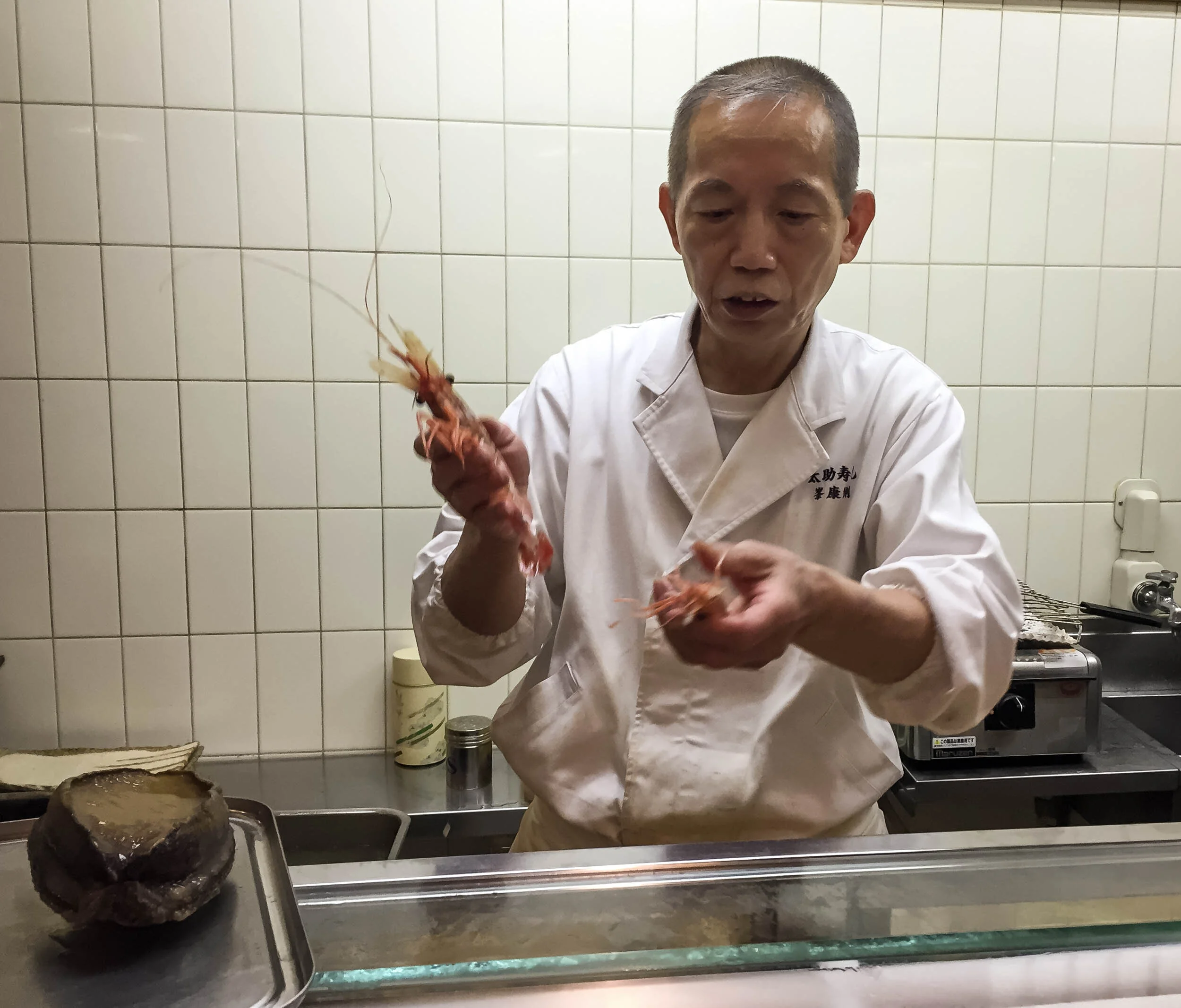 46. The master explaining about his crustaceans and his extremely choosy approach to purchasing each morning; slow-steamed abalone ("awabi") on the counter. (Tokyo, July 2015)