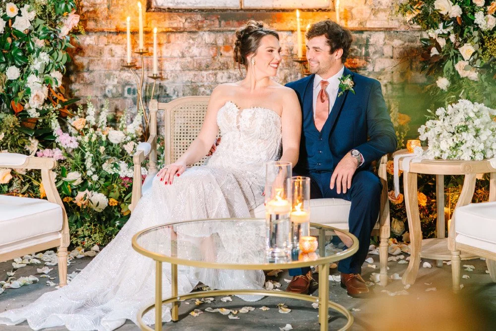 A cozy couple smiles together while sitting beneath stunning, dreamy floral arch.
