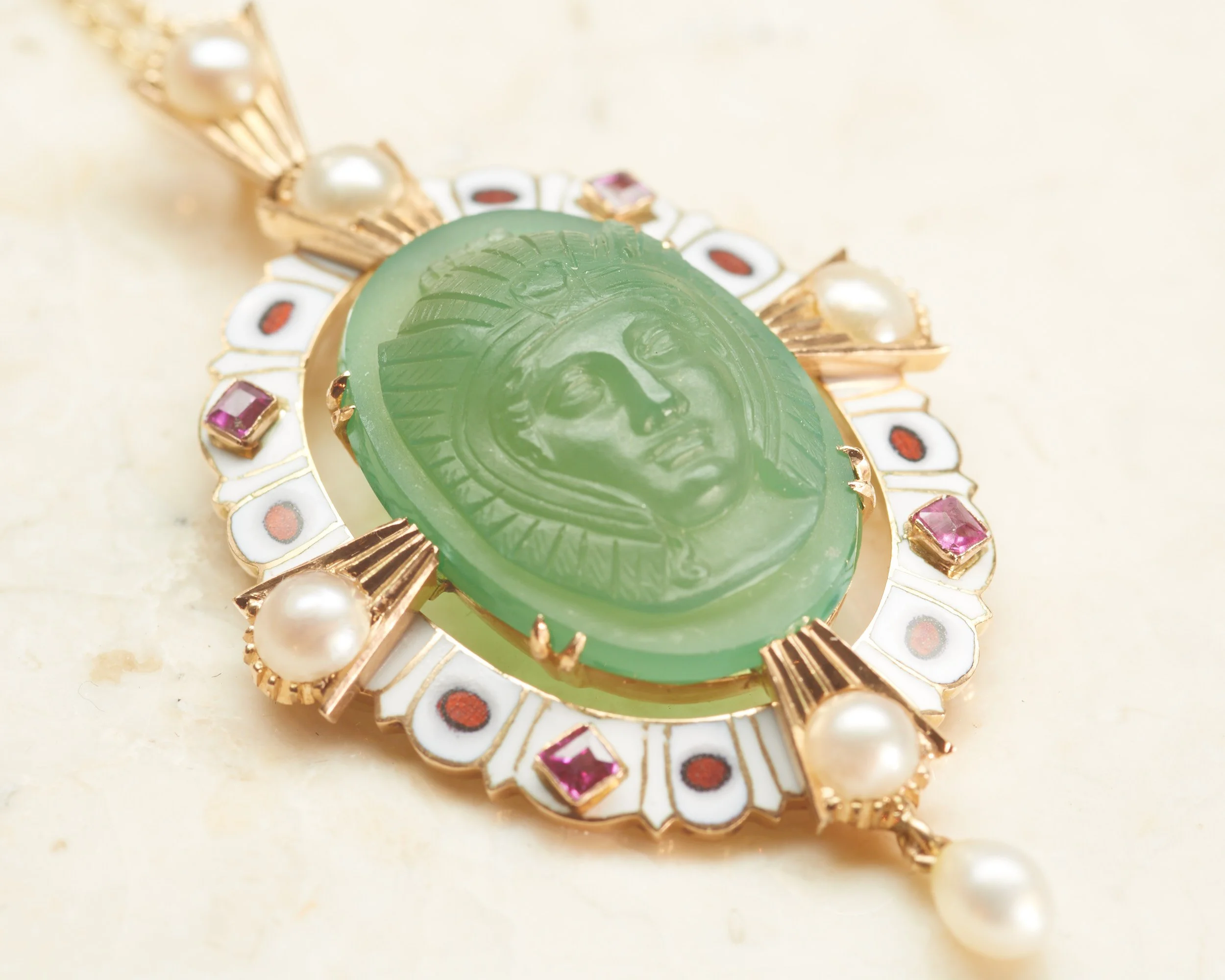 An exceptional antique Victorian 18-karat yellow gold pendant, designed in the Egyptian revival fashion of the day. Centering a carved green onyx cameo within a white enamel frame studded with four square rubies and four half-pearls, with full pearls