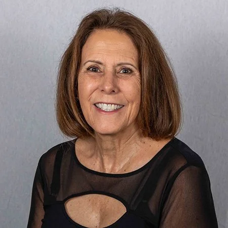 A smiling woman with shoulder-length brown hair wearing a black top with sheer fabric and cutout details, standing against a plain gray background.
