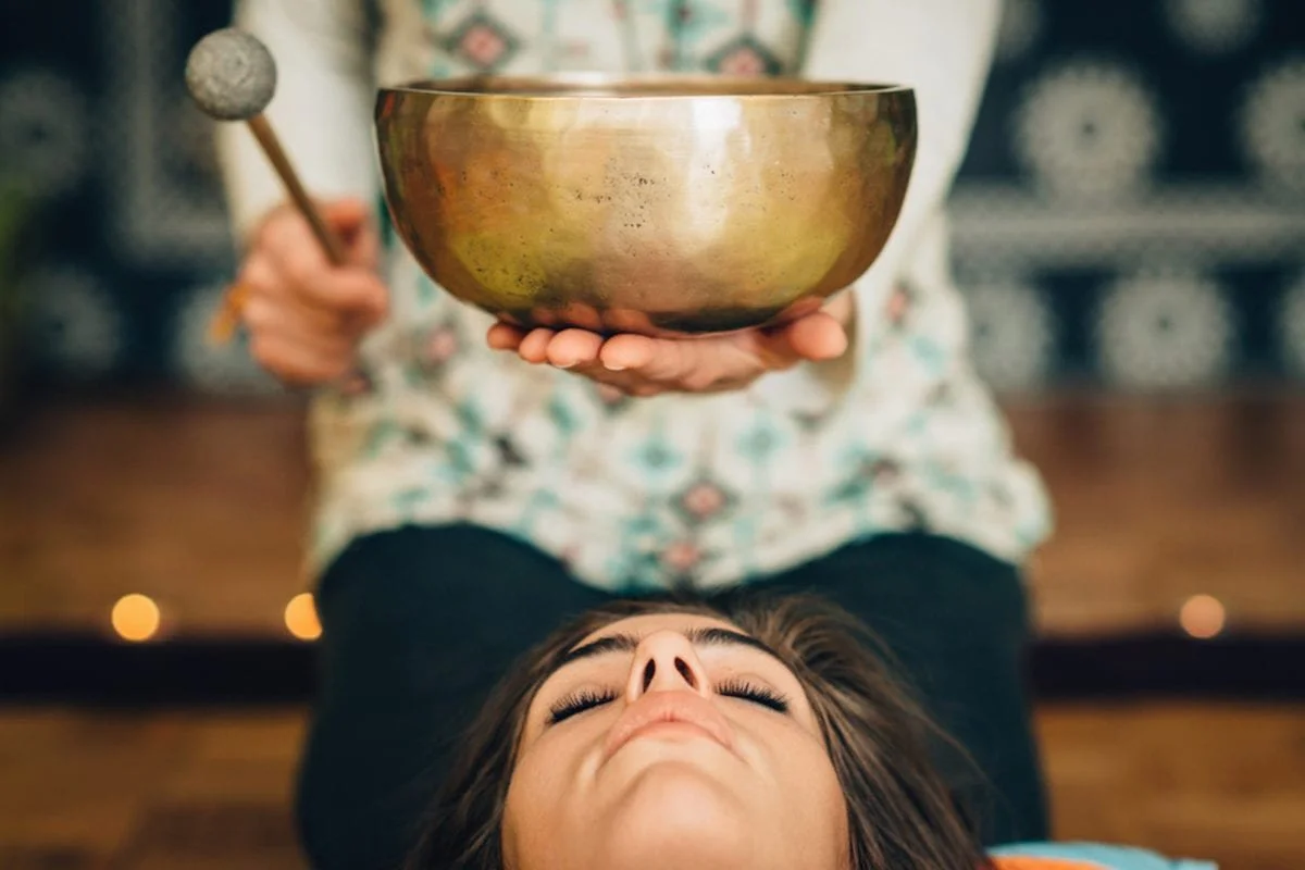 A decorative wooden table featuring a black singing bowl with an engraved symbol, a jar of white candle, a bunch of purple dried lavender, and a small potted plant in a metallic holder.