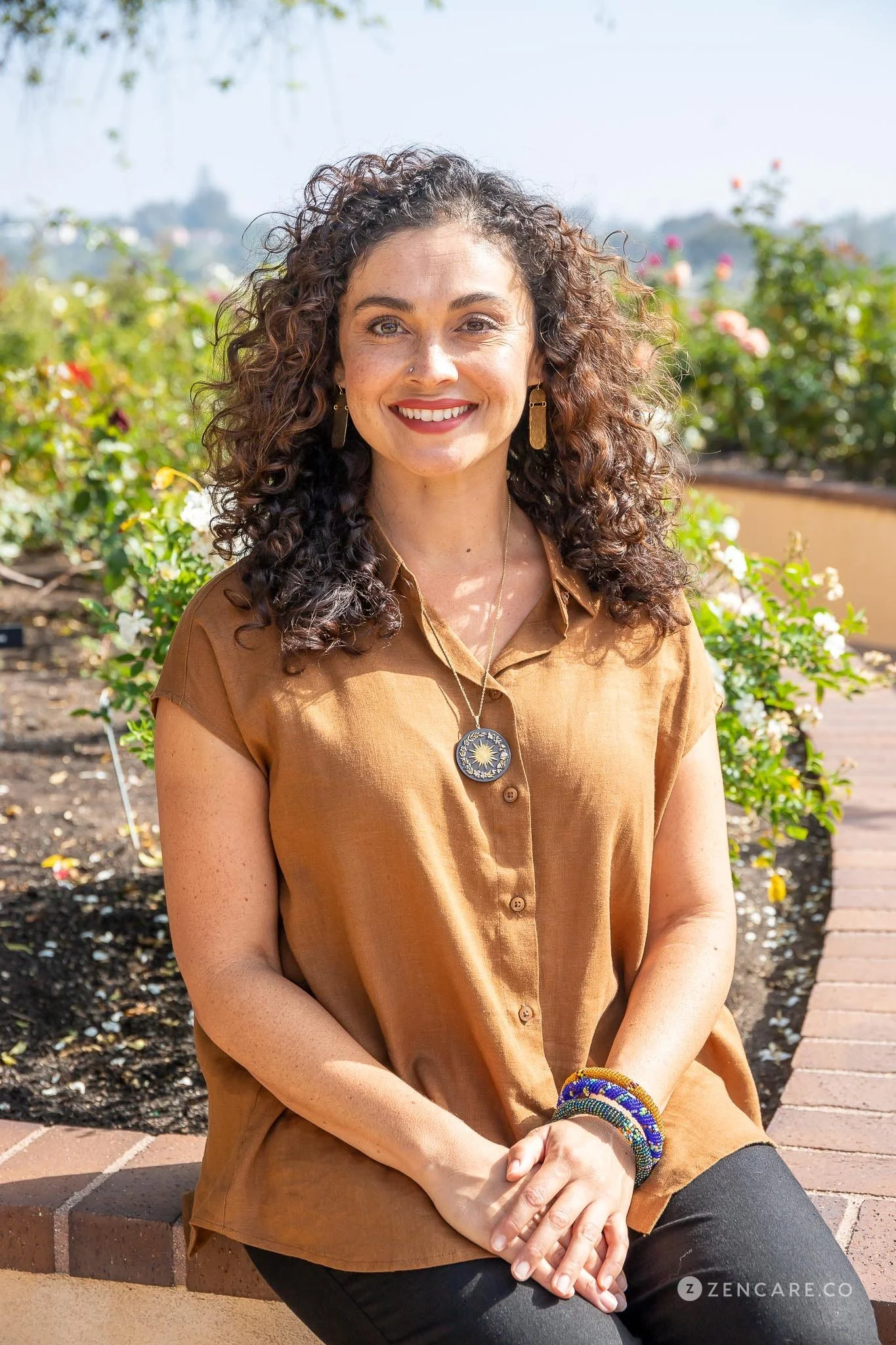A woman with curly brown hair sitting outdoors on a brick ledge, smiling, with a background of a garden with blooming flowers and a city skyline in the distance. She is wearing a brown sleeveless blouse, layered necklaces, earrings, and bracelets.