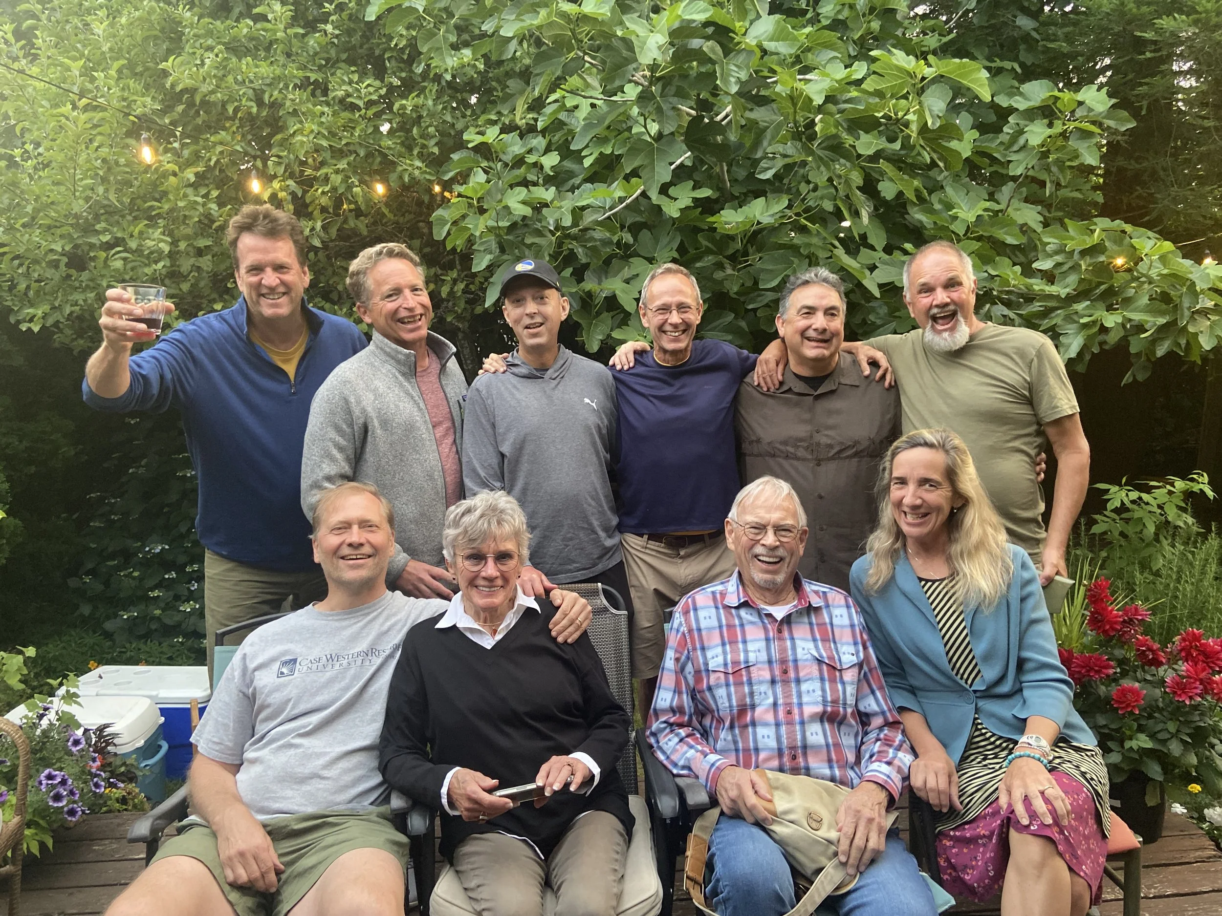 Group of people smiling and posing together outdoors in a garden, with green trees and string lights in the background.