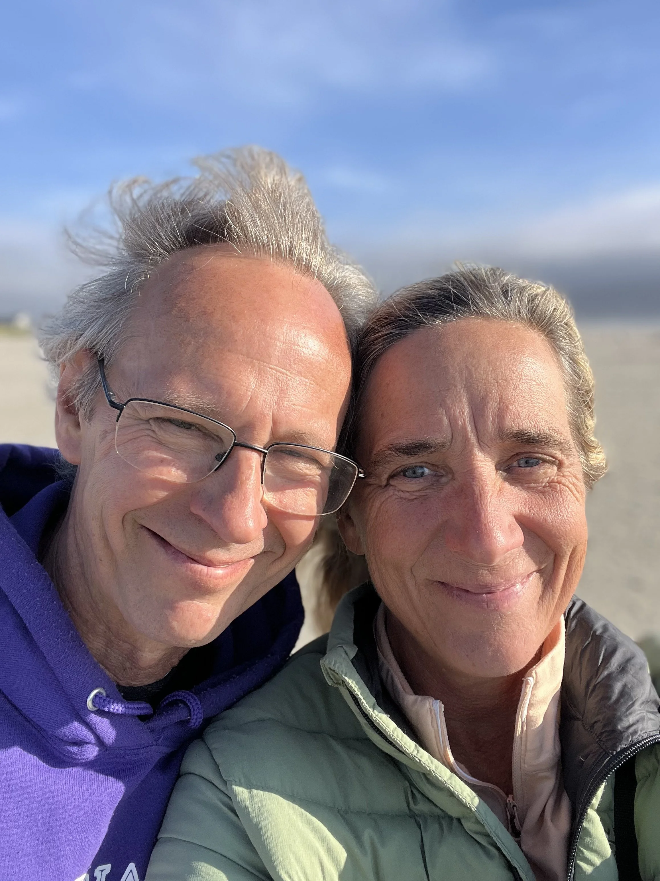Close-up of a man and woman smiling outdoors on a sunny day, with a blue sky and cloudy background.