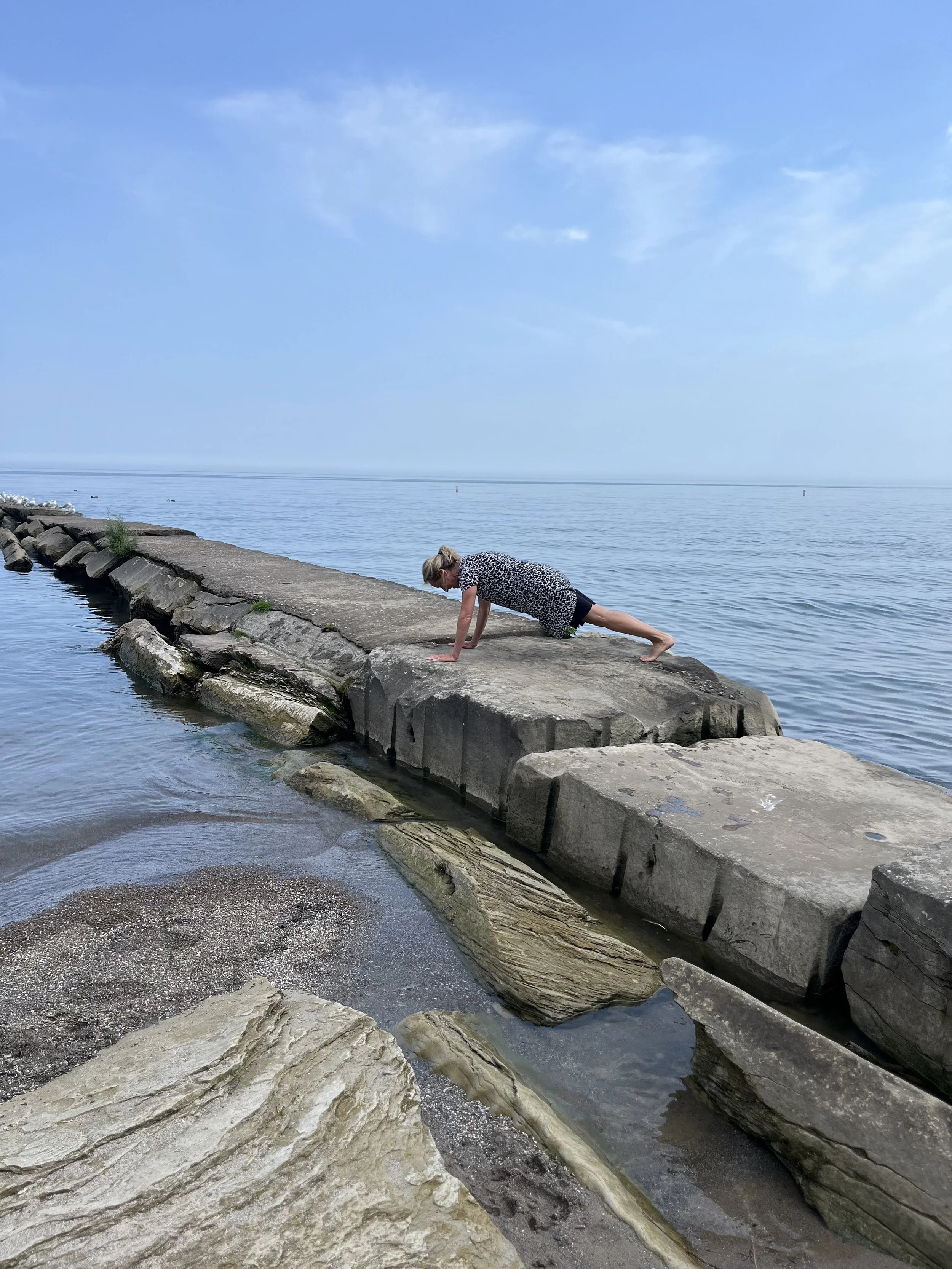 A person in a patterned dress is doing a push-up on a concrete pier by a body of water, with a cloudy sky overhead.