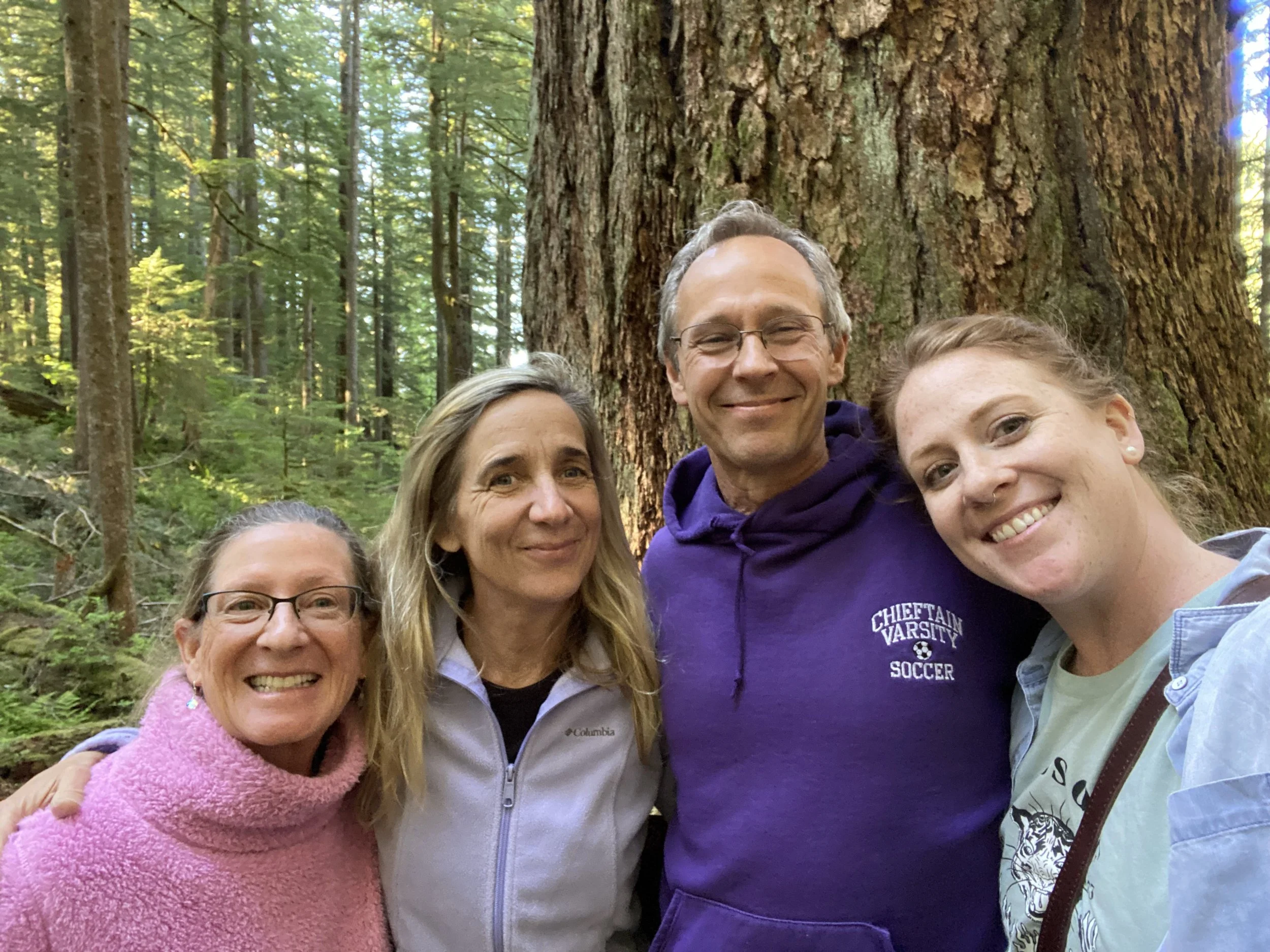 Group of five smiling people outdoors in a forest, close together, in front of a large tree with dense foliage.