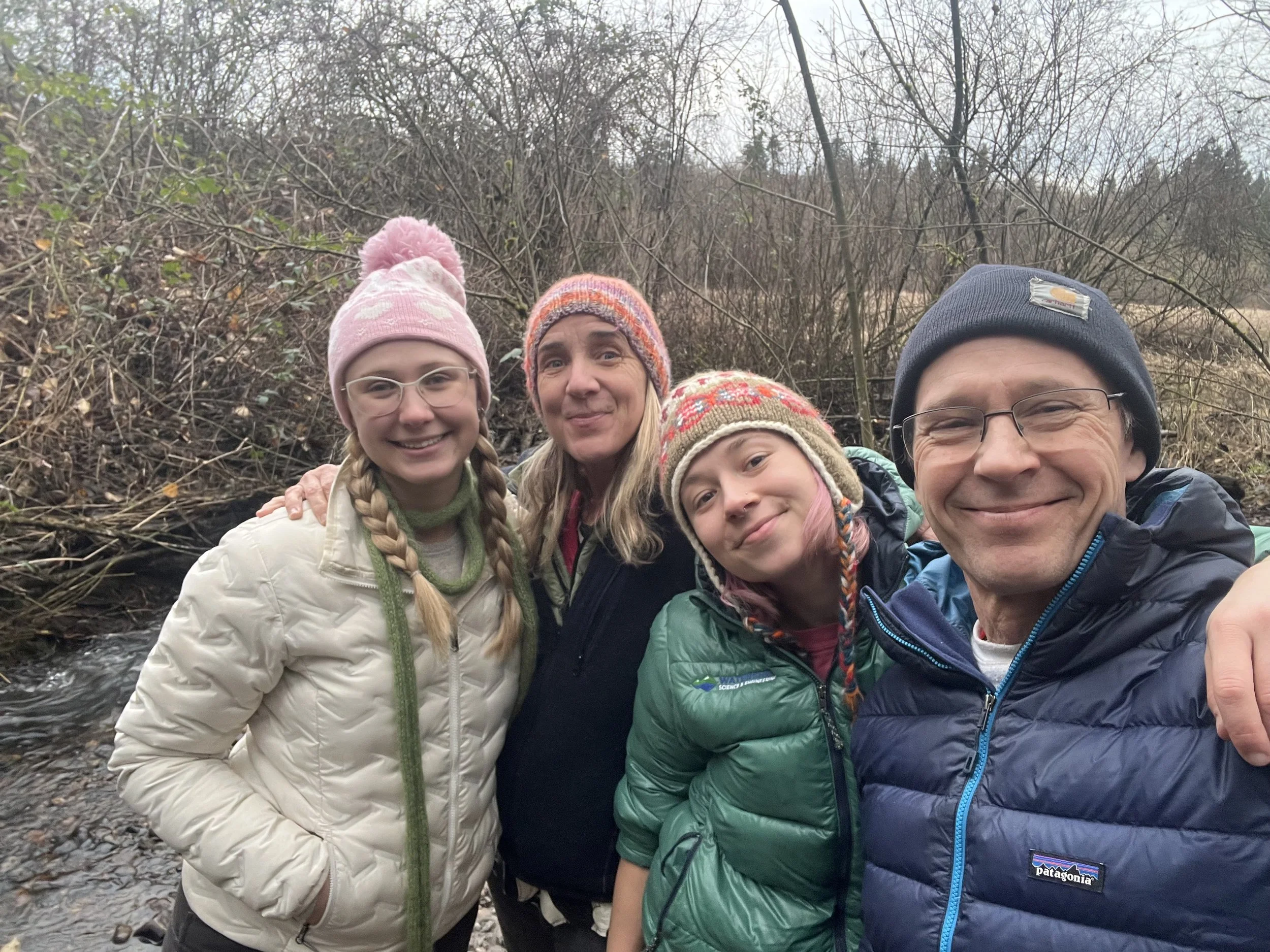 A group of five people, including children and adults, taking a selfie outdoors in a wooded area during a cloudy day, all wearing warm jackets and hats.