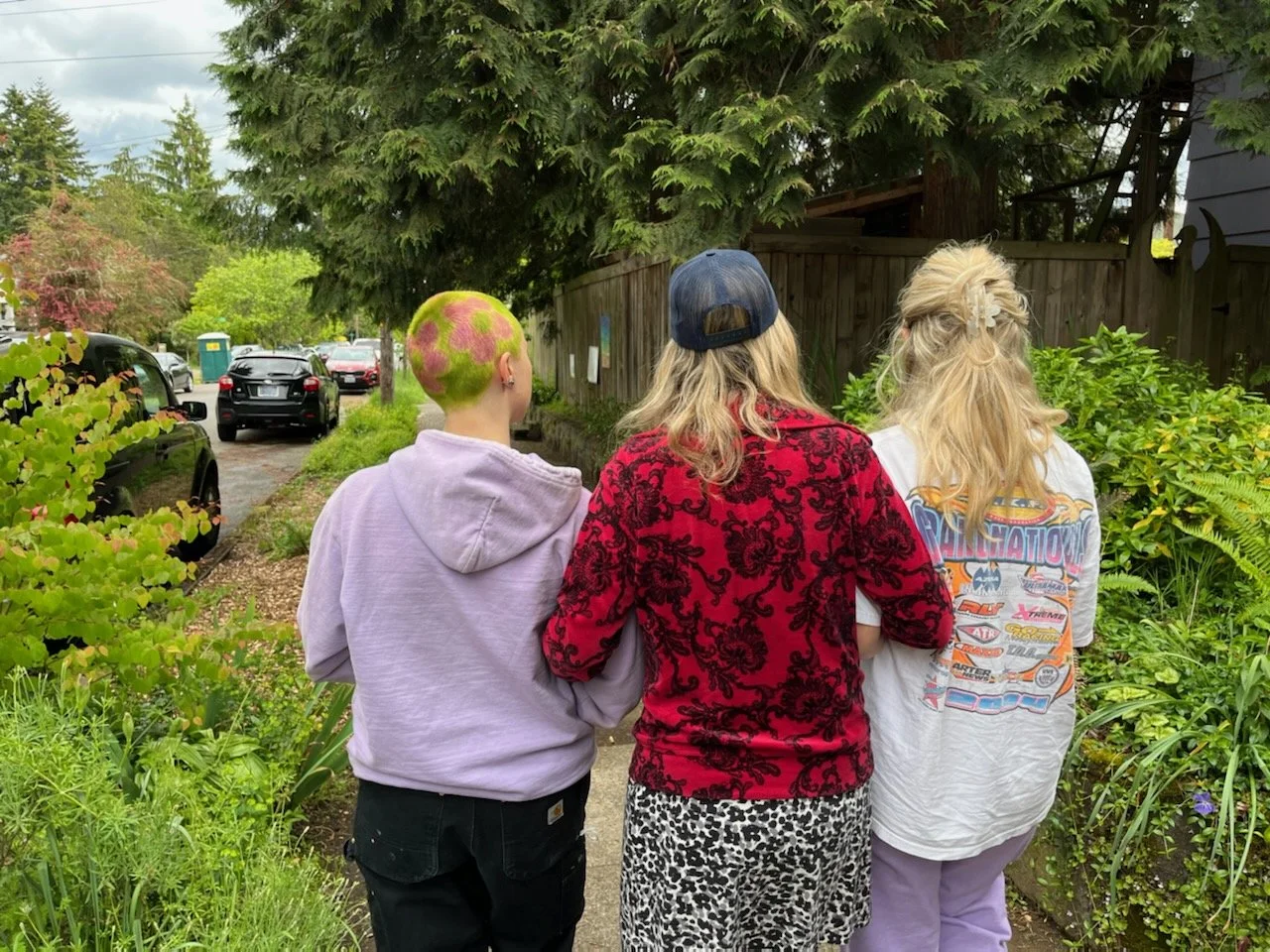 Three people walking on a sidewalk in a lush green garden with trees and plants, facing away from the camera, near parked cars and a wooden fence.