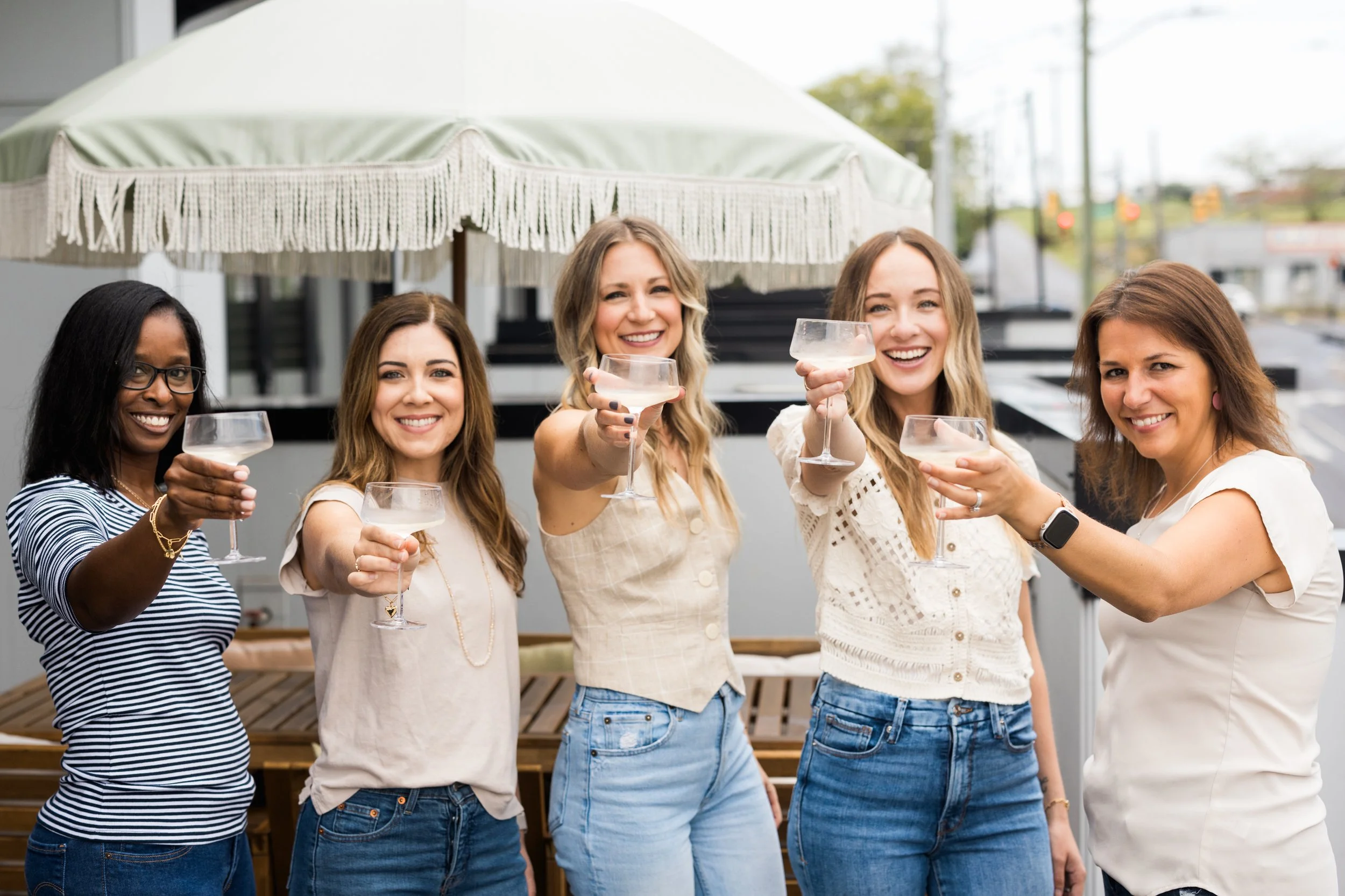 A group of five women smiling and raising glasses of champagne together, standing outdoors under a large patio umbrella.