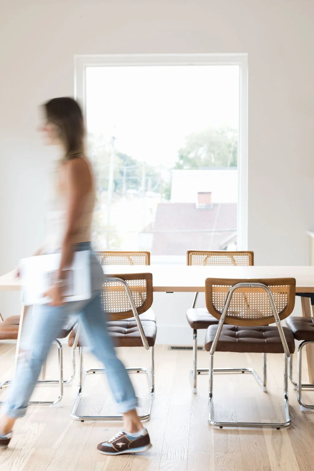 Blurred woman walking past a wooden table and chairs in a bright room with a large window.