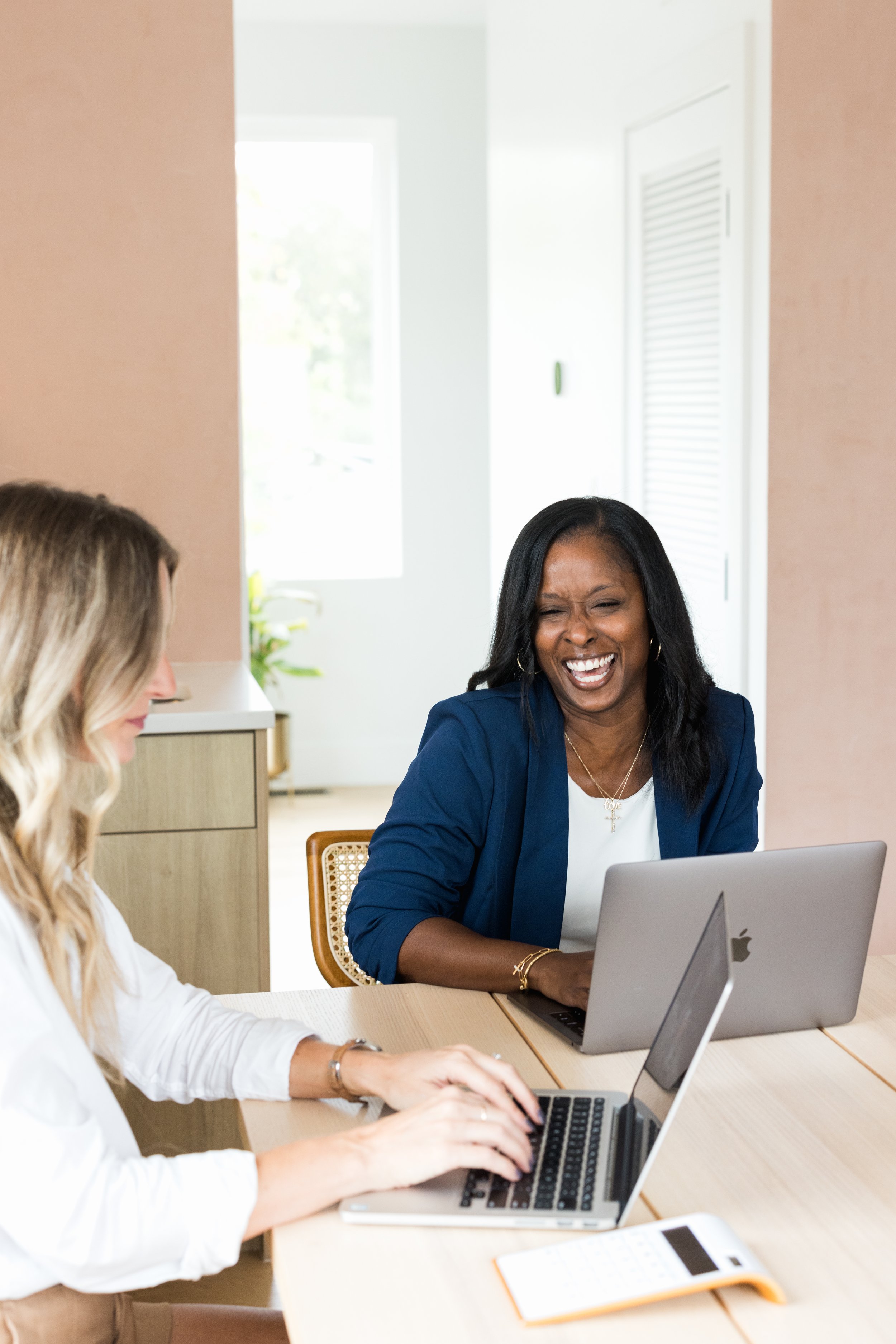 Two women sitting at a wooden table working on their laptops in a bright room, laughing and chatting.