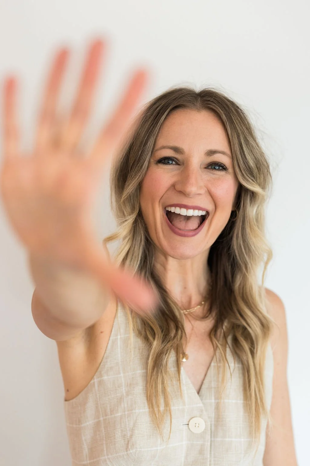 A smiling woman with long wavy blonde hair reaching out toward the camera, with her mouth open in a happy expression, wearing a sleeveless beige checkered top.