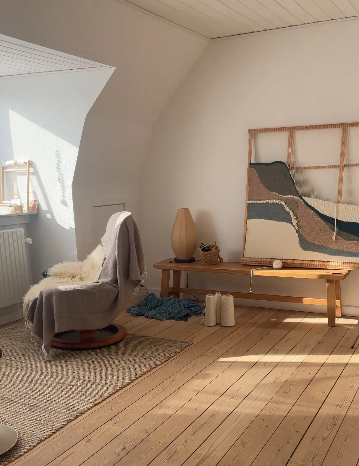 Et hyggeligt hjørne med en komfortabel stol med pude og tæppe, en træbord med en vase, en kurv og trådstrikket kunstværk på væggen, samt en radiator ved vindue, i et lyst rum med trægulv.
