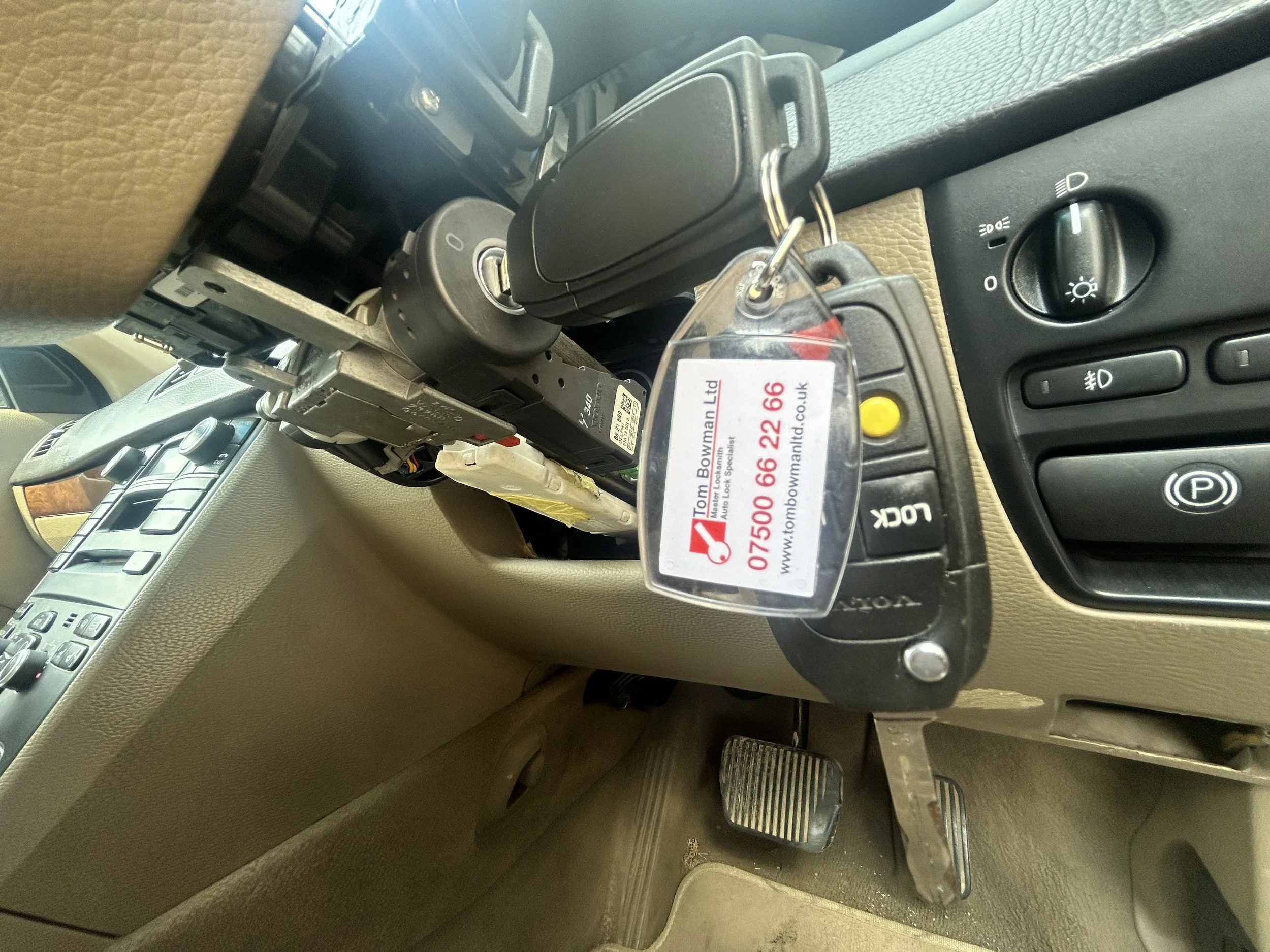 Inside a vehicle, view of the dashboard and pedals with car keys hanging from the ignition and a small business card with red writing attached to the keychain reading 'Tom Bowman Ltd, Auto Lock Specialist'