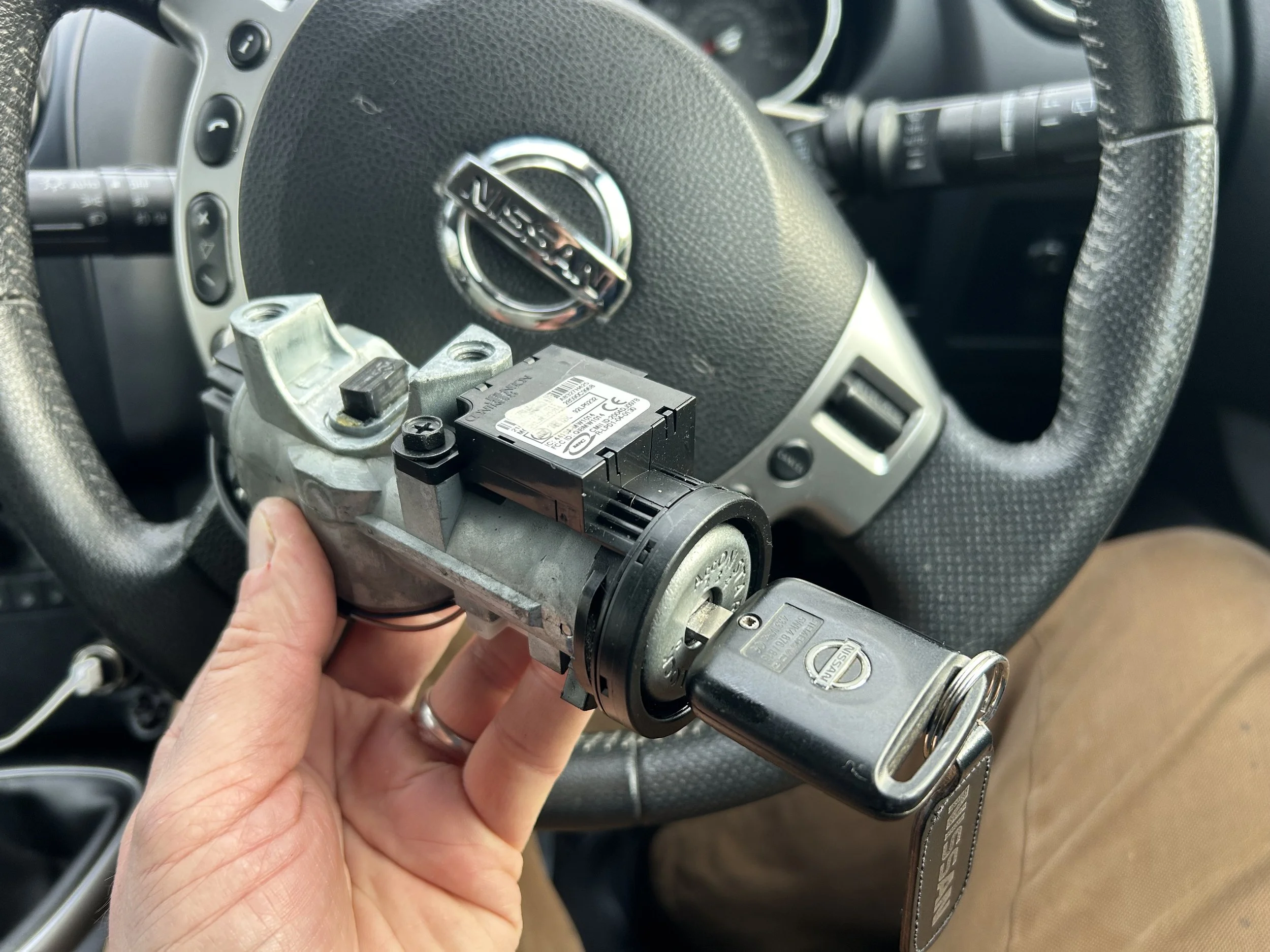 A person holding a car ignition switch module in front of a Nissan steering wheel inside a vehicle, with some dashboard controls visible.