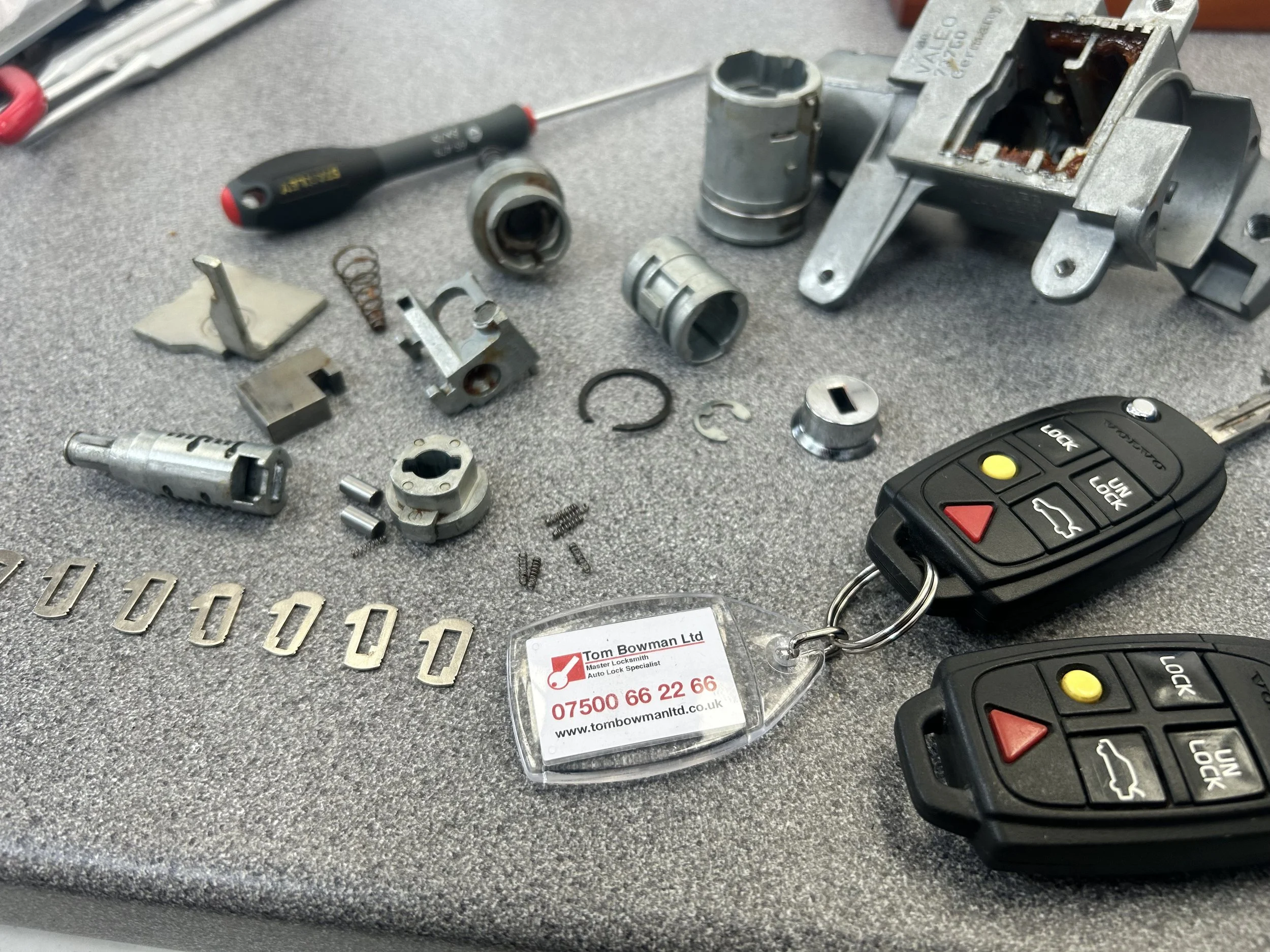 Disassembled car door lock mechanisms, small metal components, and a car remote on a grey work surface at an auto locksmith shop.
