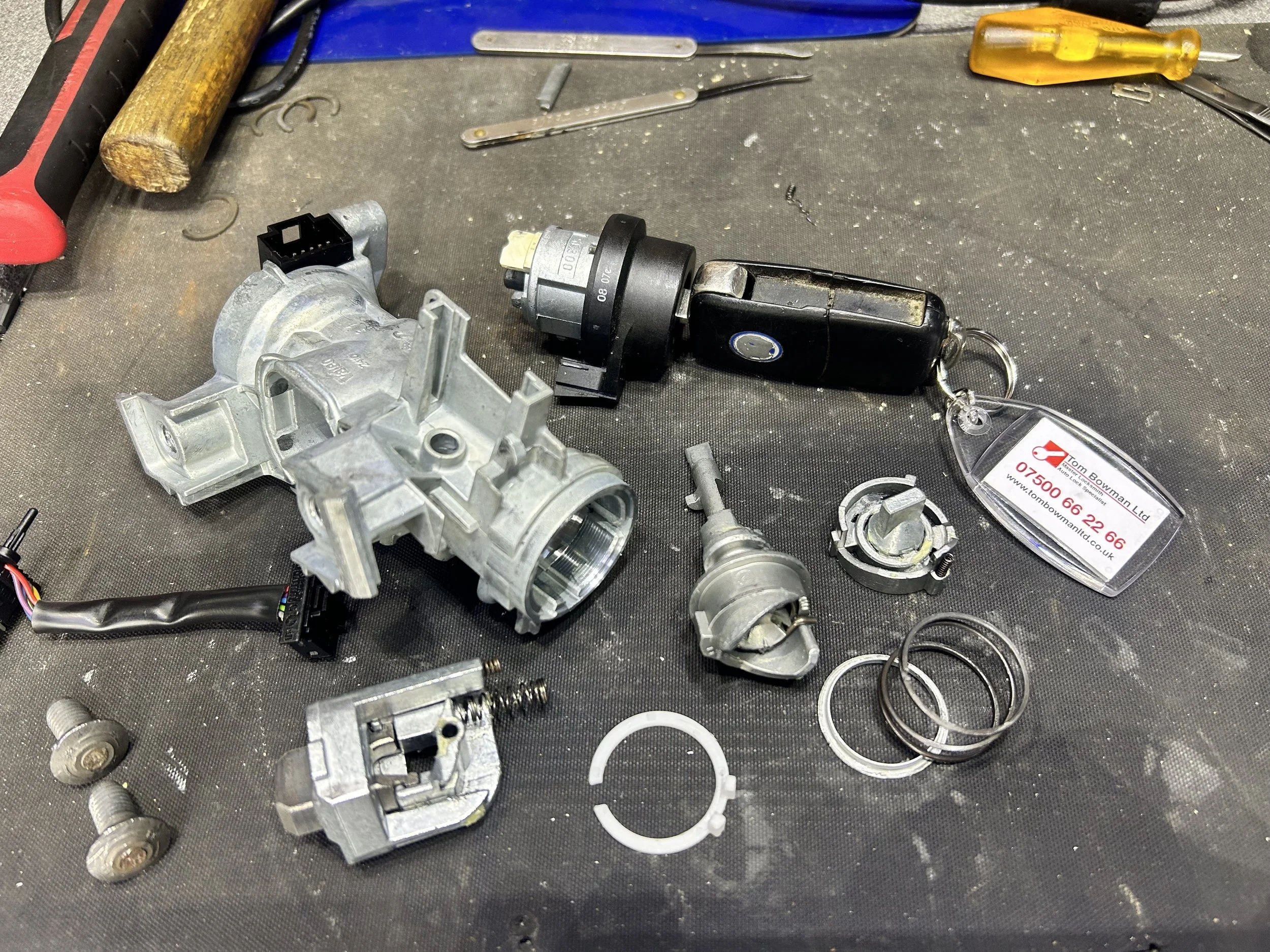 A workbench with various disassembled automotive parts, tools, and a business card. The parts include metallic components, rings, springs, and a black plastic sensor with wiring.