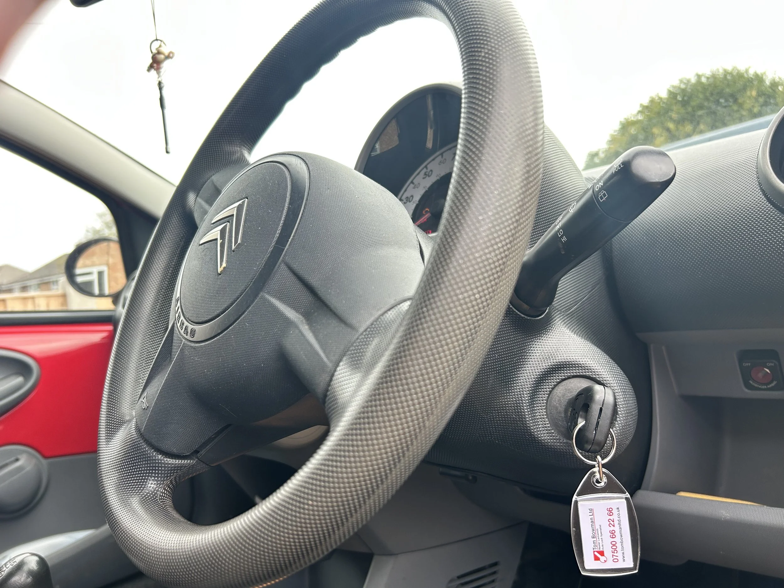 Close-up of a car's steering wheel with a textured grip and dashboard controls. A small ornament dangles from the rearview mirror, and a keychain with a business card reading 'Tom Bowman Ltd, Auto Lock Specialist' inserted in the ignition lock.