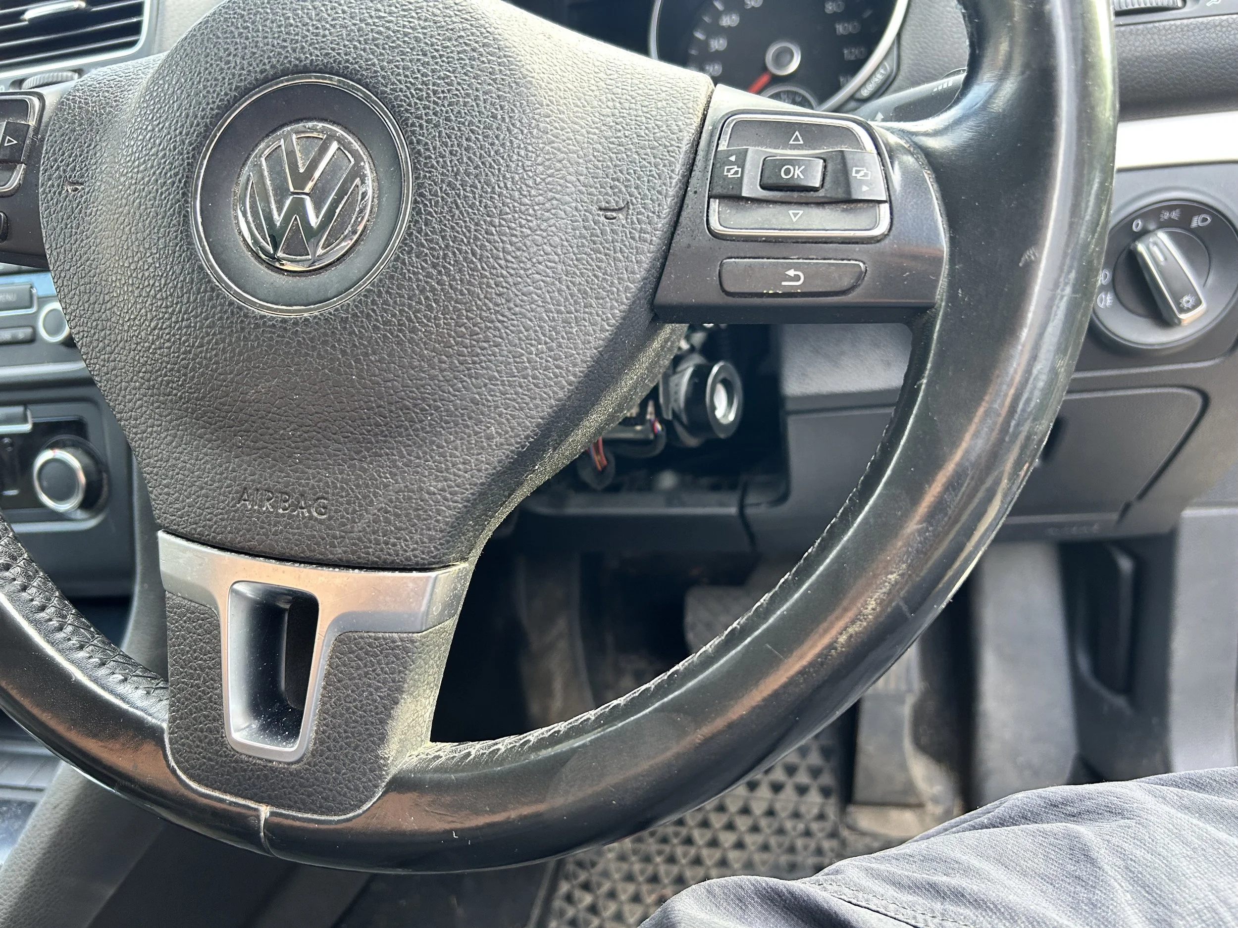 Close-up of a black Volkswagen car steering wheel with control buttons on the right side and a textured surface, with speedometer visible in the background.