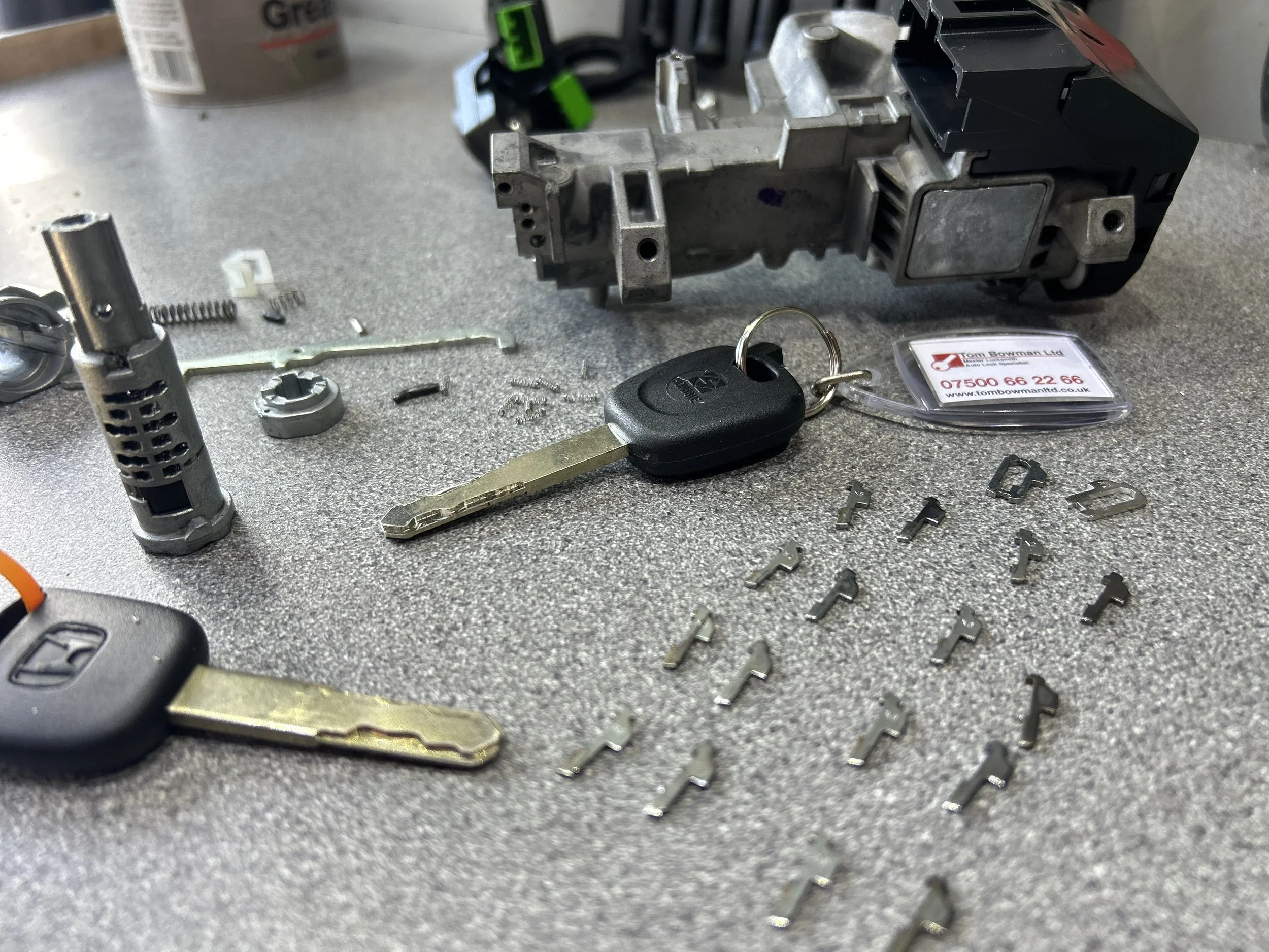 Disassembled electronic car key with black plastic, metal components, and a key blade on a gray work surface with scattered small metal parts, tools, and a blurred background.