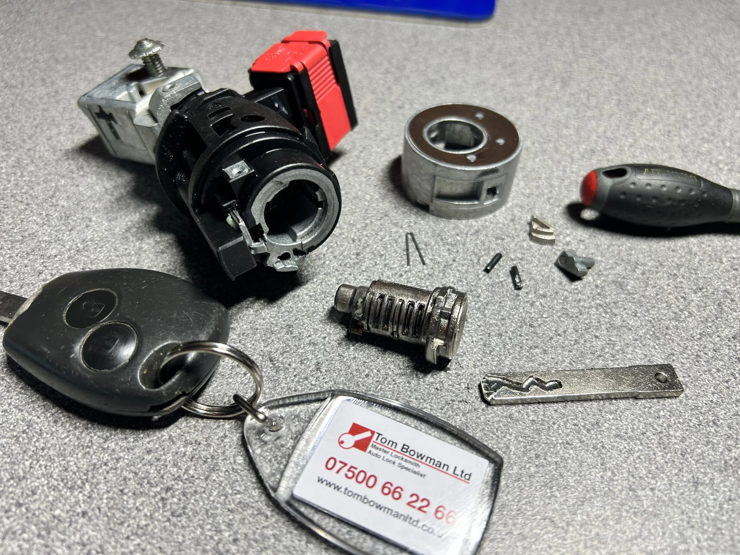Close-up of car key fob and various mechanical car lock and ignition parts on a gray surface, including a key fob with buttons, a small metal cylinder, and a business card reading 'Tom Bowman Ltd, Auto Lock Specialist' with contact information.