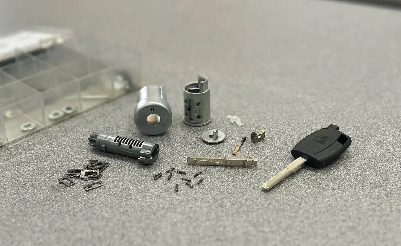 Disassembled car key and lock components on a gray surface, including a black key fob, various metal parts, and small screws.