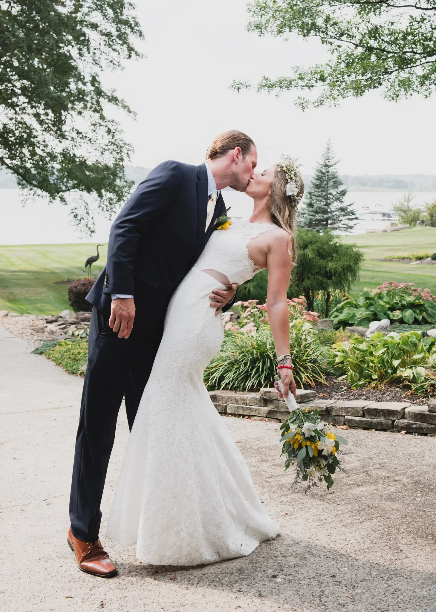 A newlywed couple sharing a kiss outdoors, the bride in a lace white wedding gown holding a bouquet, the groom in a dark suit with a sunflower boutonniere. They are in a garden setting with trees, flowers, and a body of water in the background.