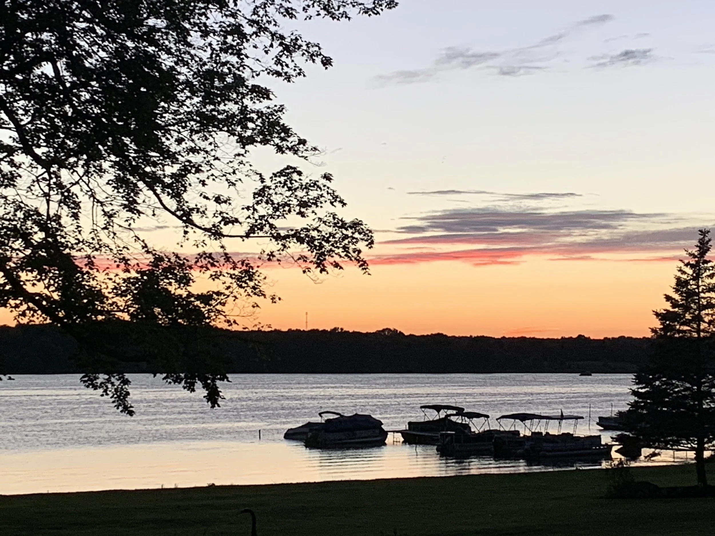 A lakeside scene at sunset with a colorful sky, silhouetted trees, and boats moored along the shore.
