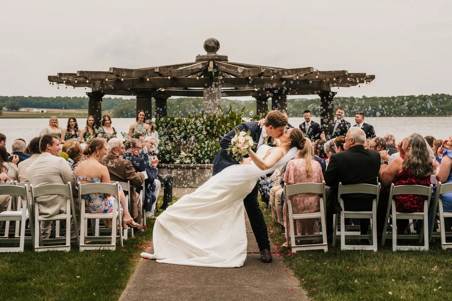 A newlywed couple shares a kiss during their outdoor wedding ceremony by the water, with guests seated nearby and a group of bridesmaids and groomsmen in the background under a wooden pergola.