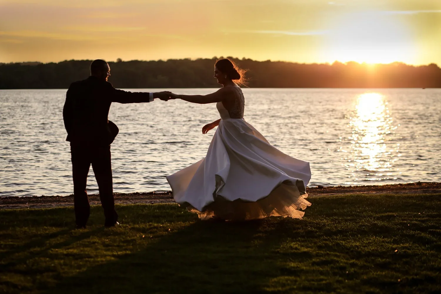 A silhouette of a couple dancing by the water at sunset, with the woman in a wedding dress and the man in a suit.