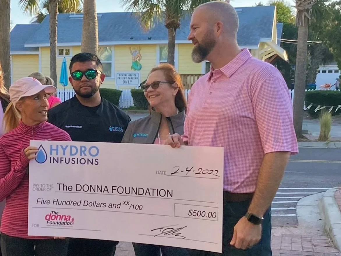 A group of people outdoors holding a large donation check for $500 made out to The Donna Foundation from Hydro Infusions, dated February 4, 2022. They are standing on a brick pathway lined with palm trees. A few other people are visible in the backgr