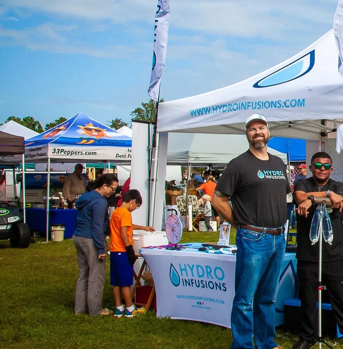 A group of people standing in front of a "Hydro Infusions" booth at an outdoor event. The booth features a canopy with flags and a table displaying promotional items. People are walking and browsing nearby stalls under a partly cloudy sky.