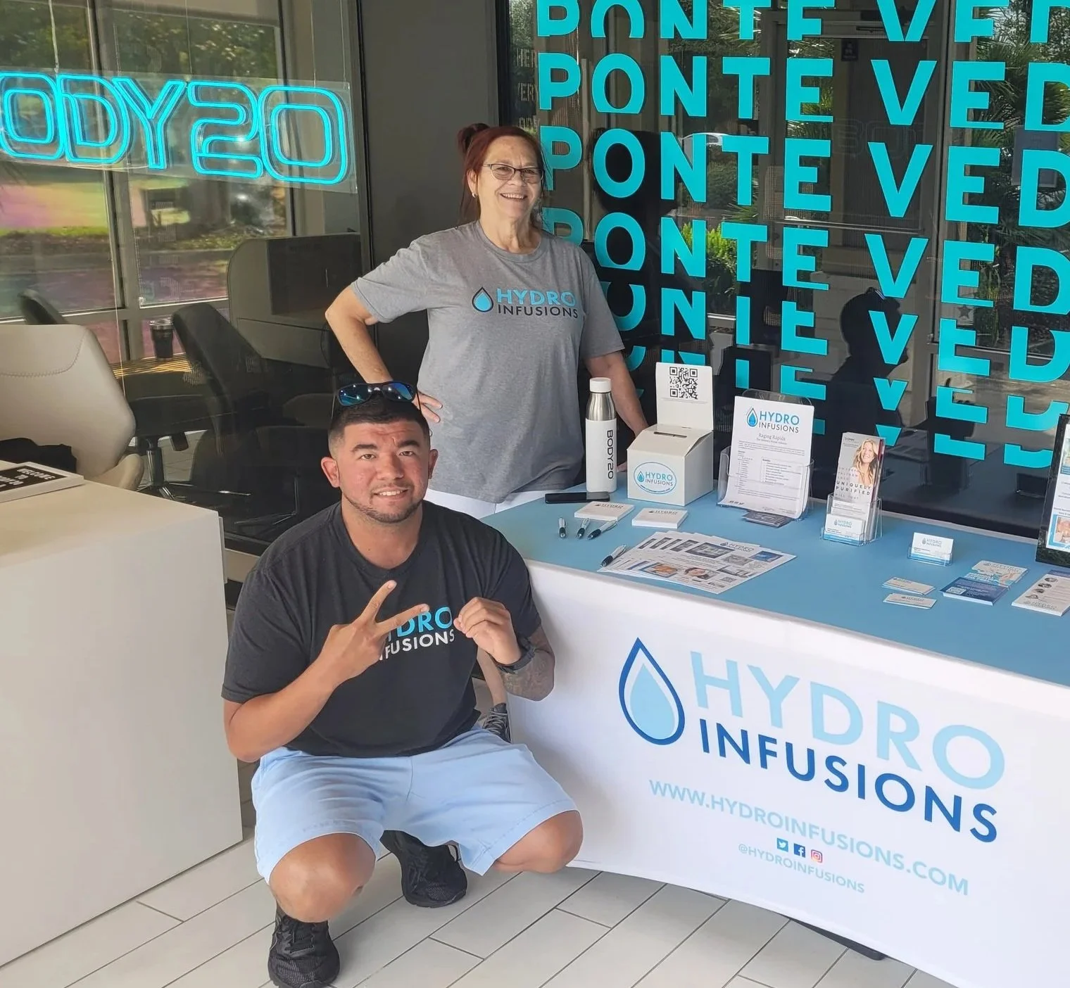 Three people at a promotional booth for Hydro Infusions, with promotional items displayed on a table. The backdrop has "Ponte Vedra" in blue text.