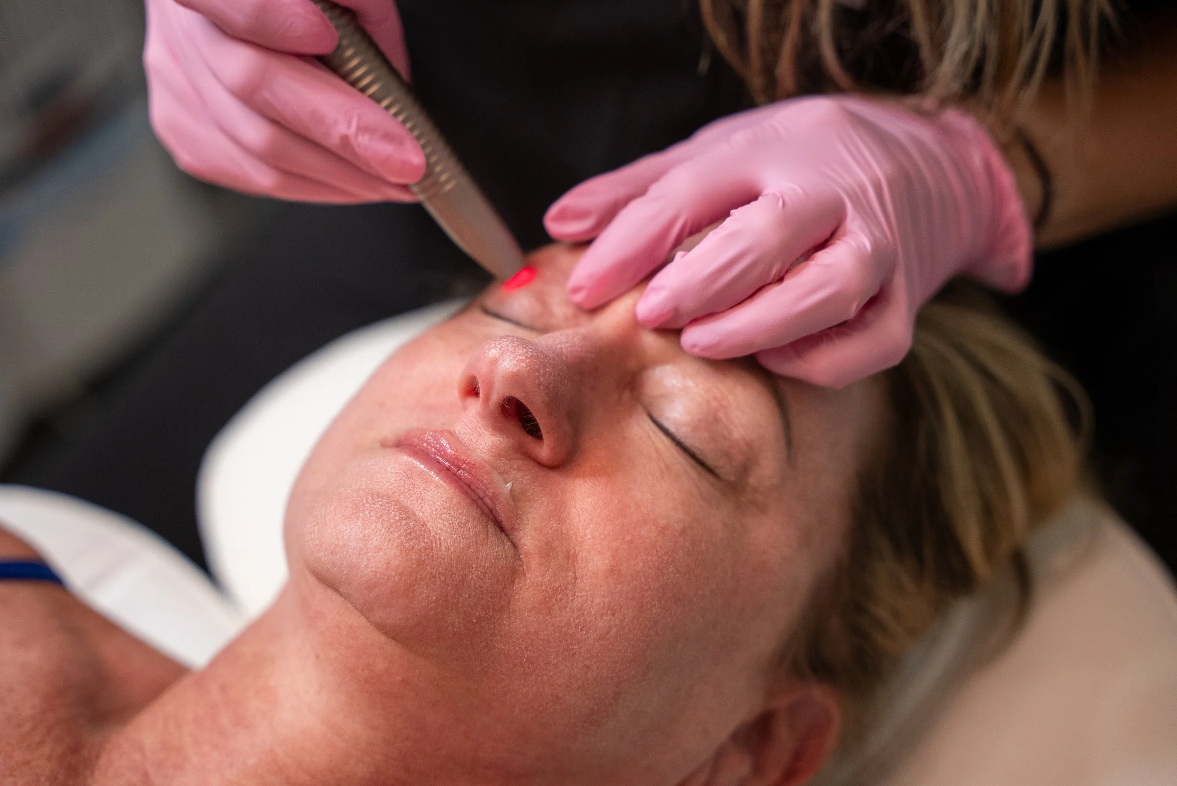 Woman receiving a facelift treatment from another woman wearing pink gloves.