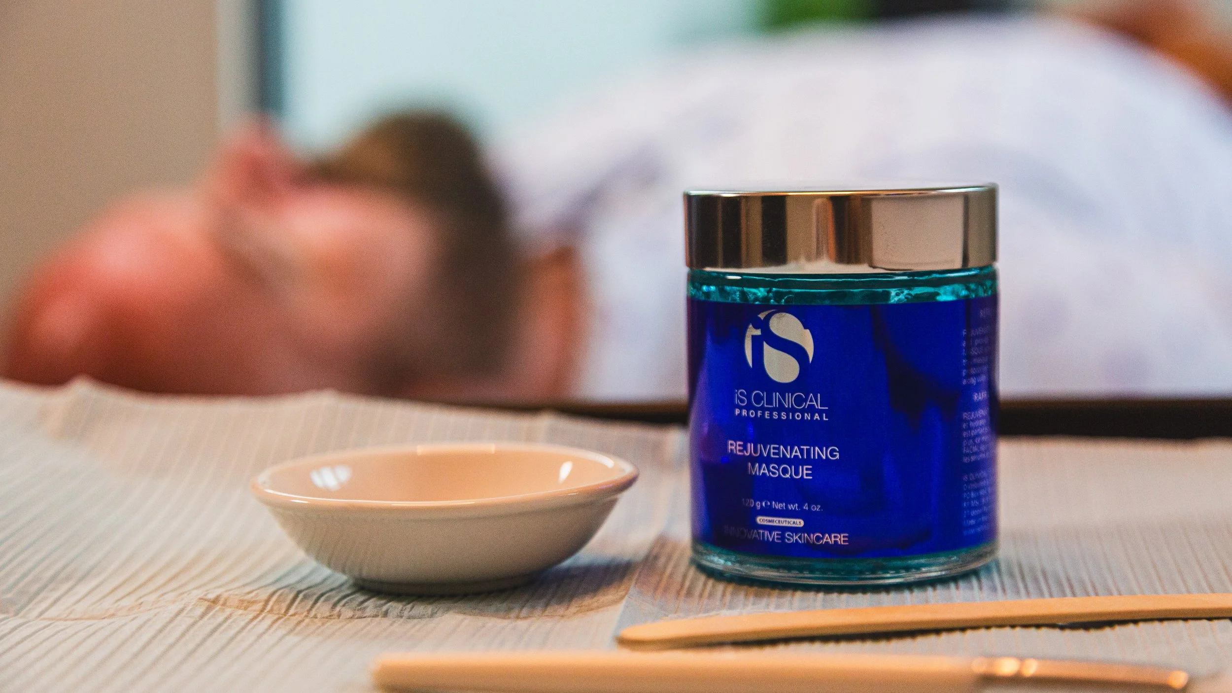iS Clinical Rejuvenating Masque jar on treatment table with client receiving facial in background at HYDRO Med Spa.