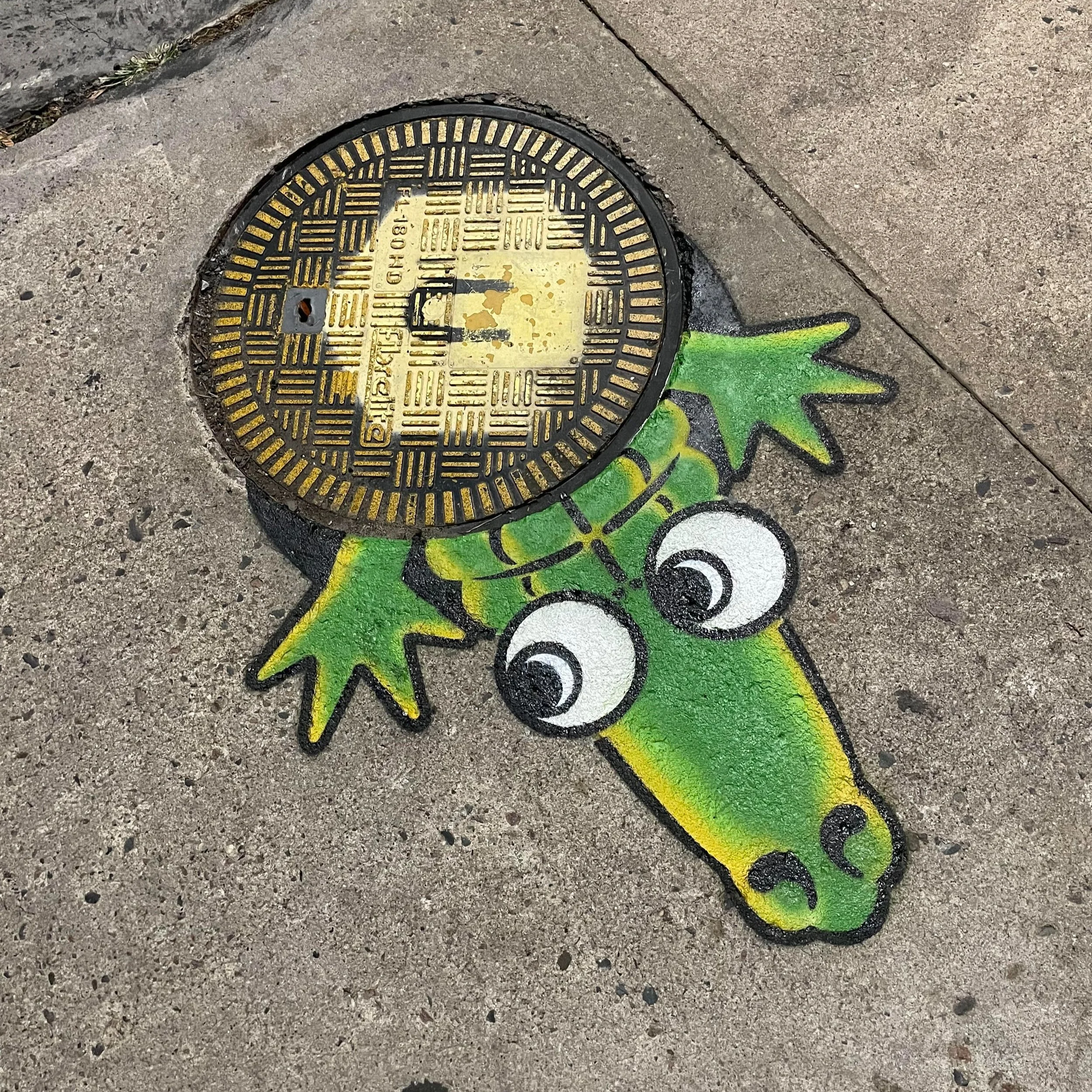 Colorful sidewalk art designed as a cartoon alligator with big eyes and a smile, painted around a manhole cover on the concrete sidewalk.