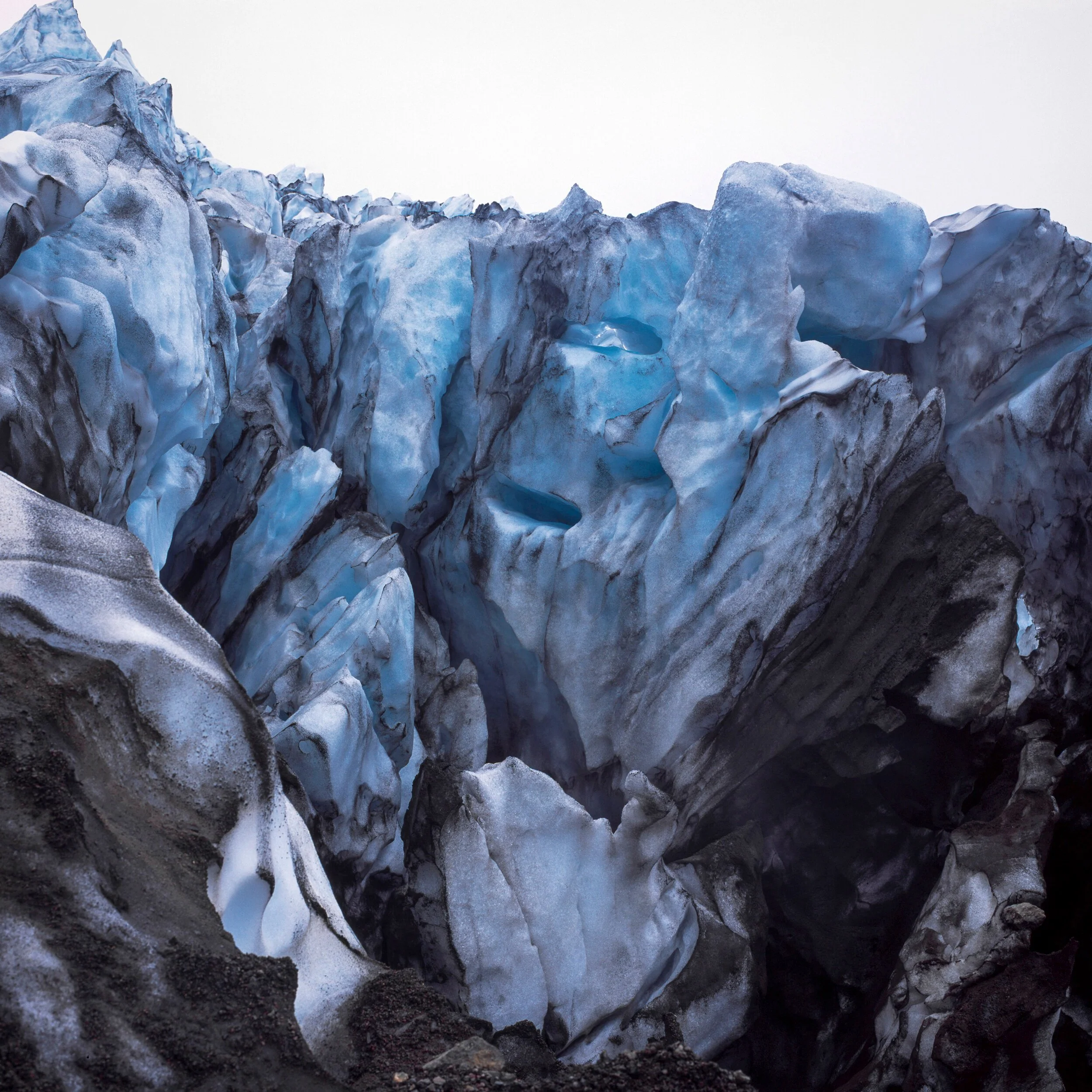 Glacier, towering wall of fractured blue ice and dark sediment at glacier edge, deep crevasses and smooth white ice against pale overcast sky, glacier in Iceland