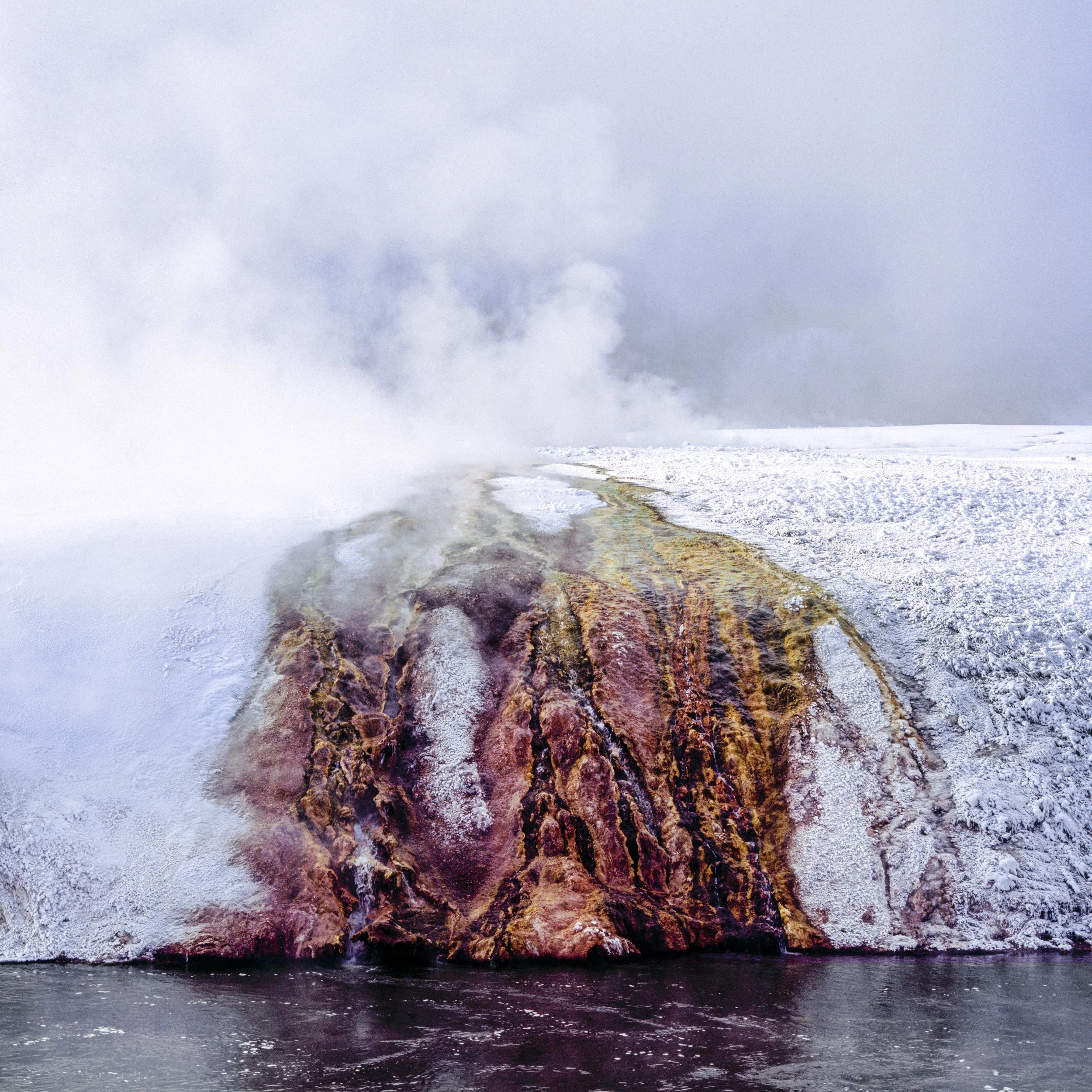 Bleed, steaming hot spring mound streaked with rust red and orange mineral runoff, edged by snow and dark river water below, Yellowstone National Park in winter