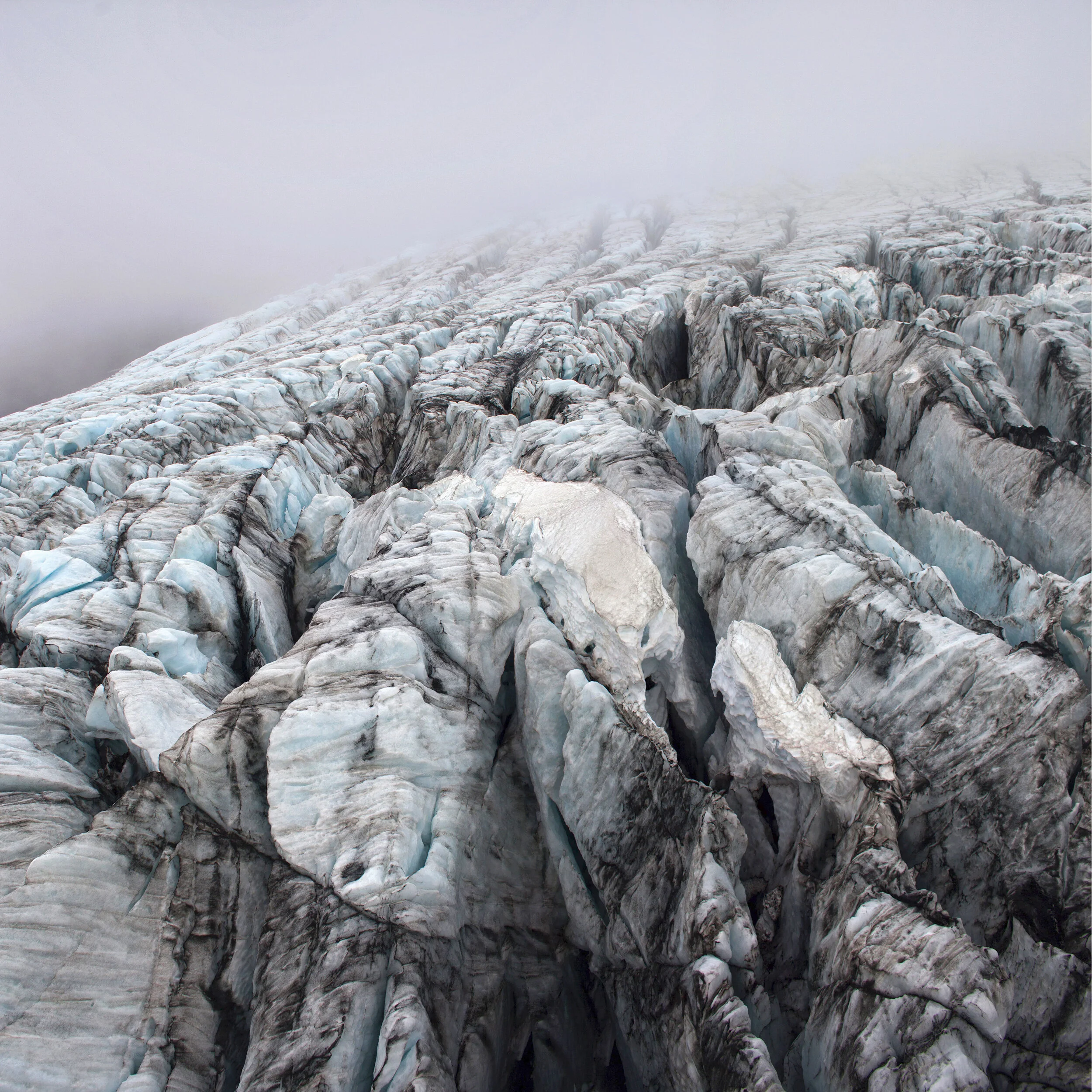 The Edge, crevassed glacier surface of blue ice and dark sediment ridges fading into white mist, jagged frozen terrain seen from above, glacier landscape in Iceland