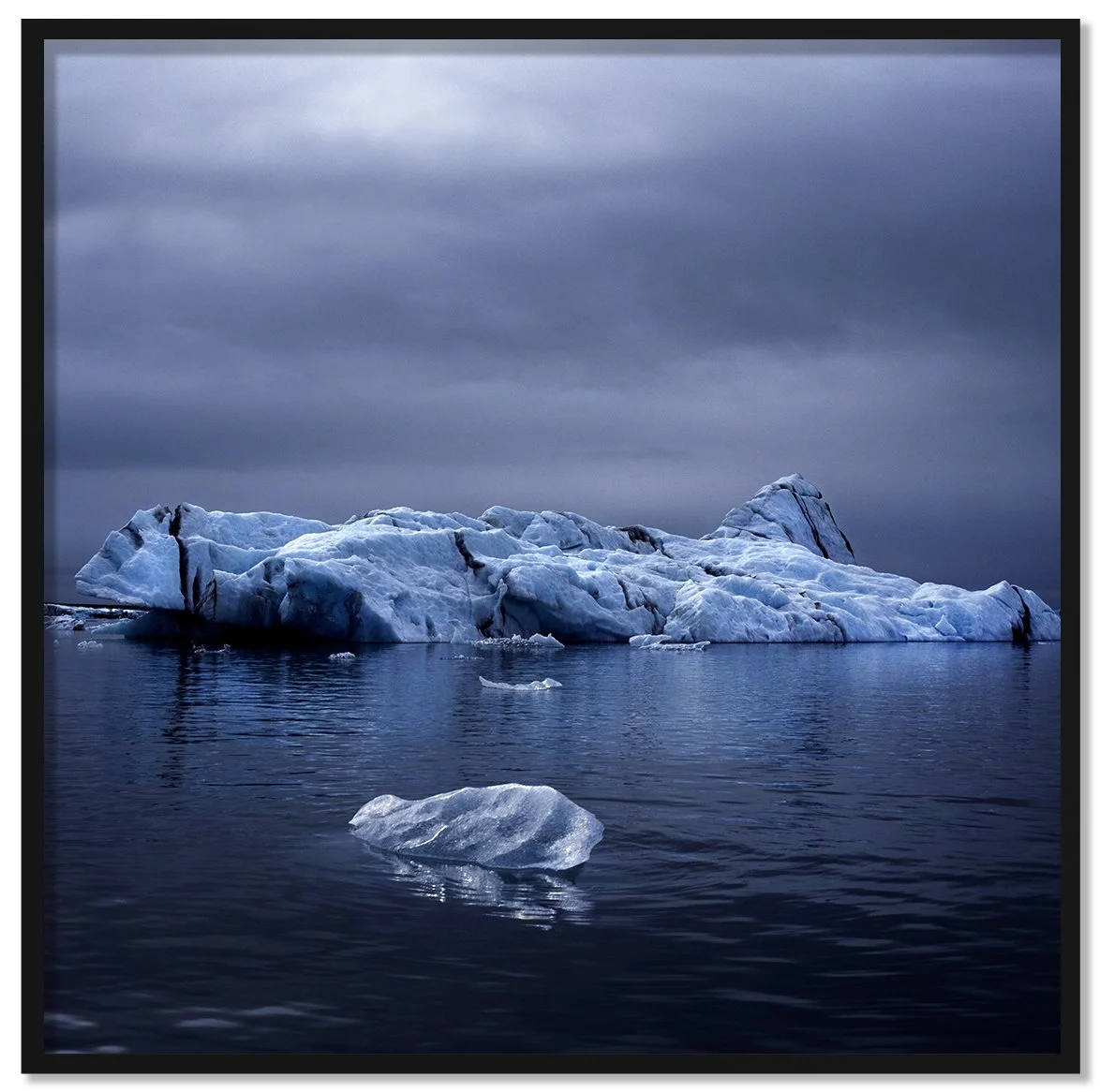 Drifter framed fine art print by Luca Marziale, blue and white iceberg floating on dark still water under heavy grey sky, small ice fragment in foreground, Iceland