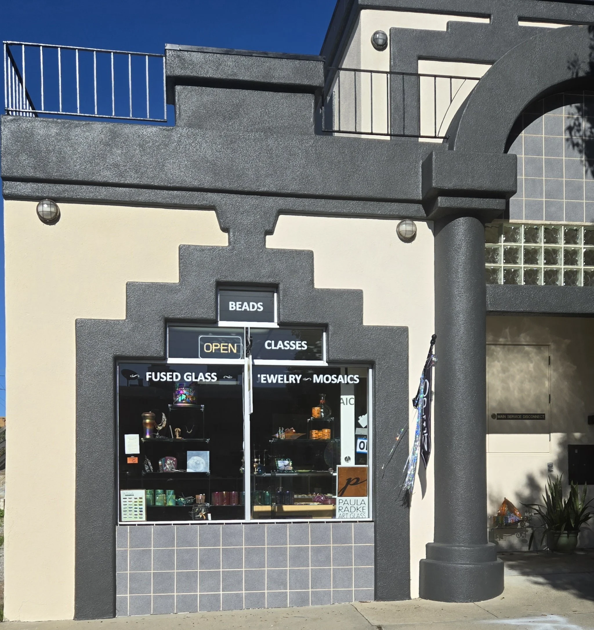 storefront with signs that read Beads, Fused Glass, Jewelry, Mosaics, and Classes, with a glass window display of art glass, beads, and jewelry, outside of a building with a beige wall and dark gray trim.