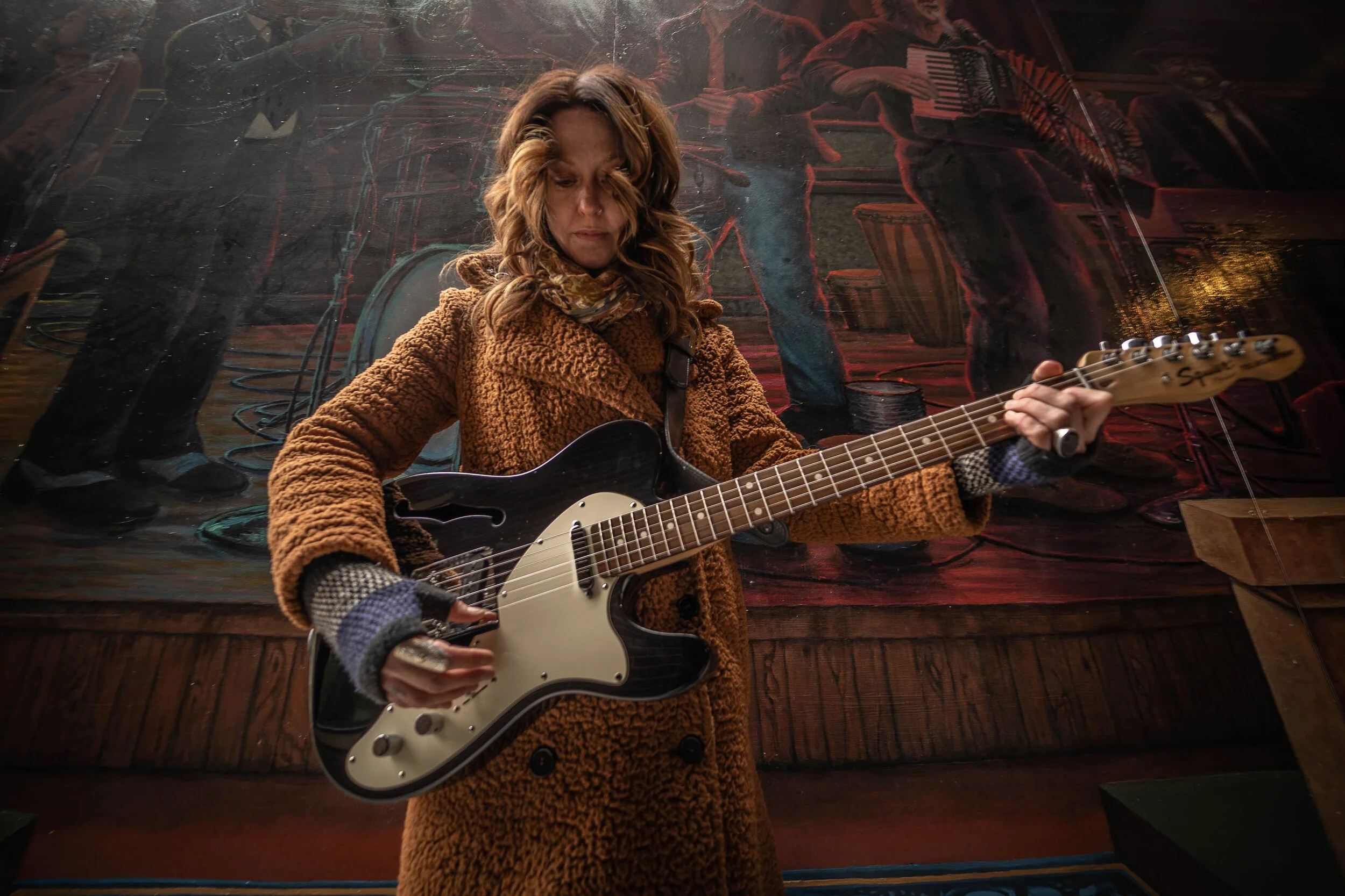 A woman with brown, wavy hair wearing a brown coat with fuzzy texture, playing a black and white electric guitar inside, with a mural showing musicians in the background.